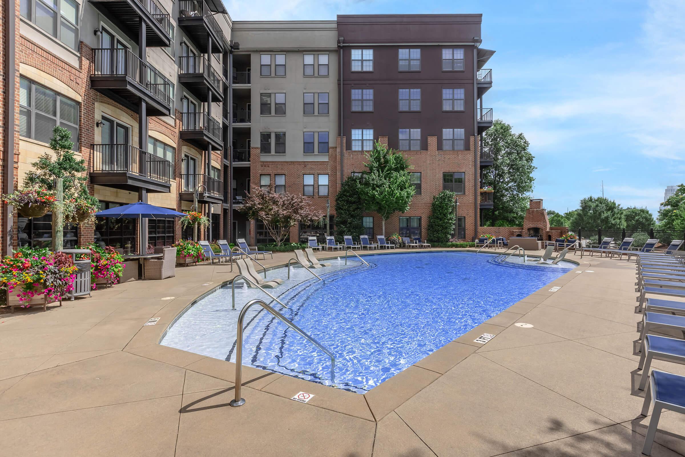 A well-maintained outdoor swimming pool surrounded by lounge chairs, with flowering plants and trees nearby. The pool area is part of a modern apartment complex featuring brick and contemporary architecture, with clear blue skies above.