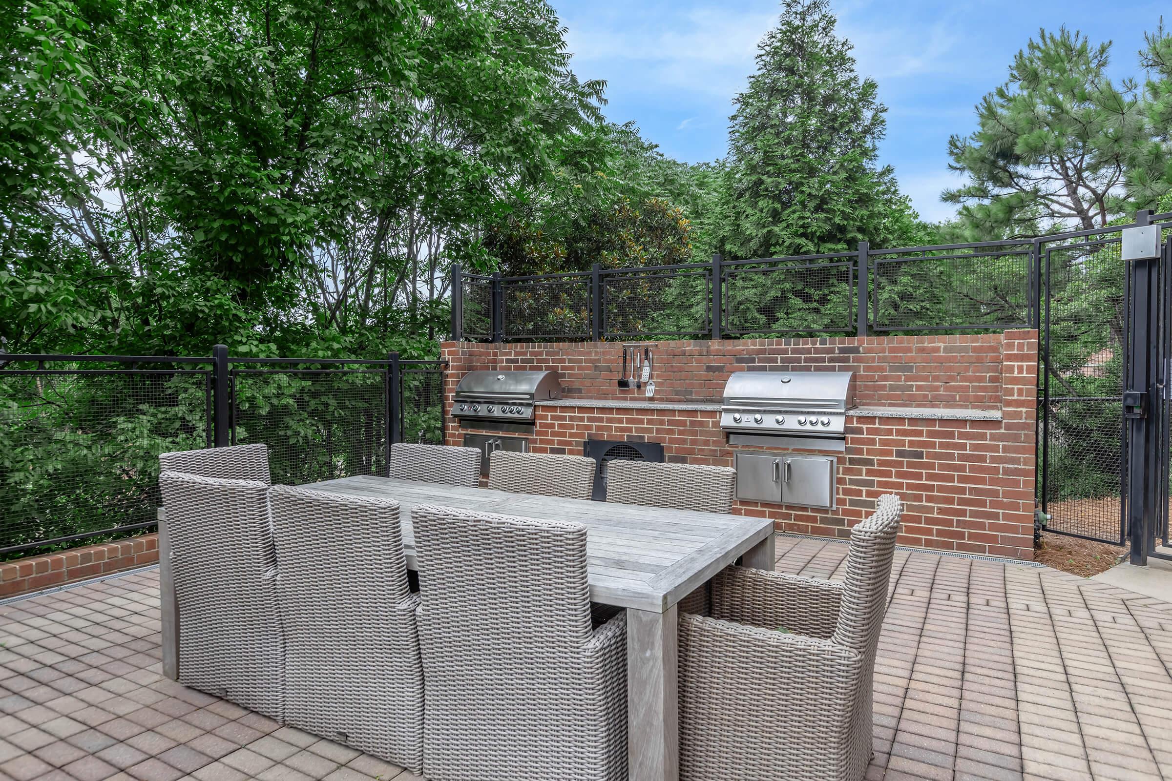 Outdoor patio featuring a modern stone dining table surrounded by eight wicker chairs. A built-in barbecue grill is mounted on a brick wall, with lush green trees in the background and a clear blue sky above. The area is enclosed by a black fence, creating a private and inviting space for outdoor dining and relaxation.