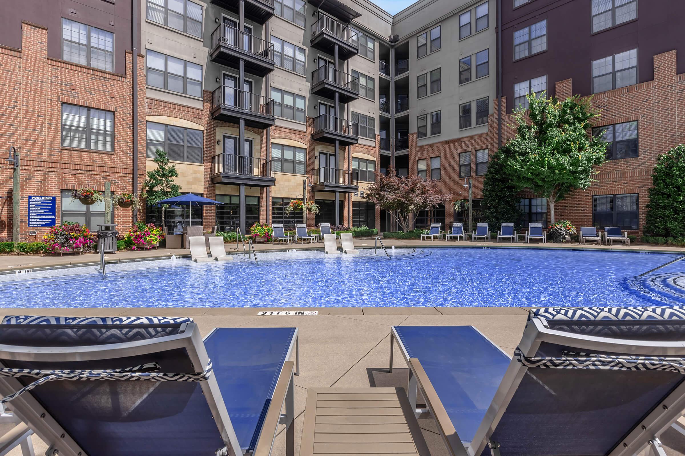 A spacious outdoor pool surrounded by lounge chairs and colorful flowers. Modern apartment buildings are visible in the background, creating a vibrant community atmosphere. The pool area is well-maintained with clear blue water, inviting residents to relax and enjoy the sun.