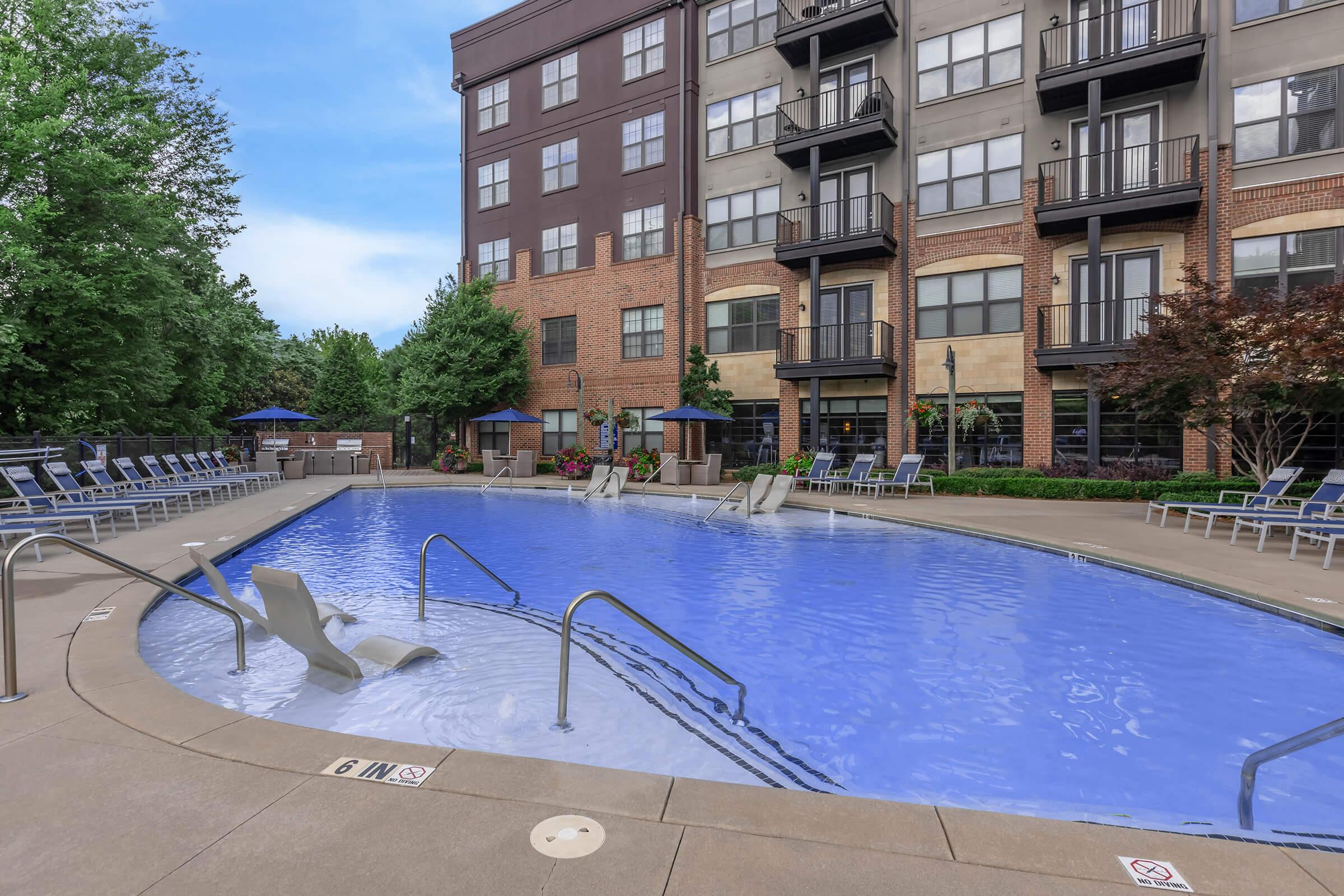 A spacious outdoor swimming pool surrounded by lounge chairs, with landscaped greenery and apartment buildings in the background. The pool features a gentle slope and accessible seating, creating a relaxing atmosphere for residents. Clear blue skies complete the scene.