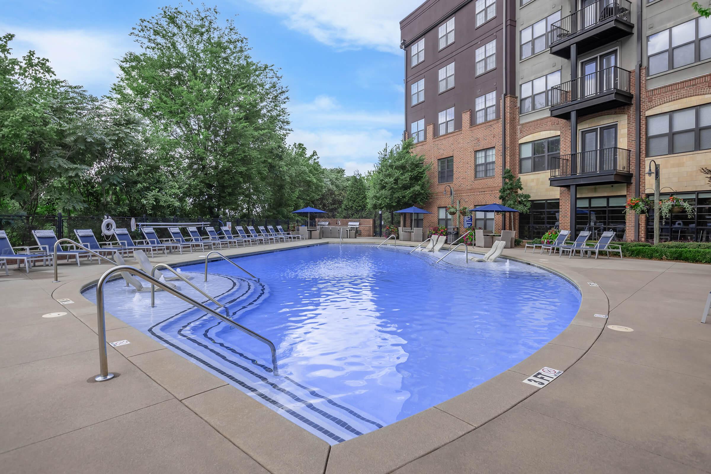 A clear swimming pool surrounded by lounge chairs and greenery, located outside a multi-story building. The pool features steps for entry, and there are umbrellas for shade. The scene is set under a clear blue sky, creating a relaxing atmosphere.