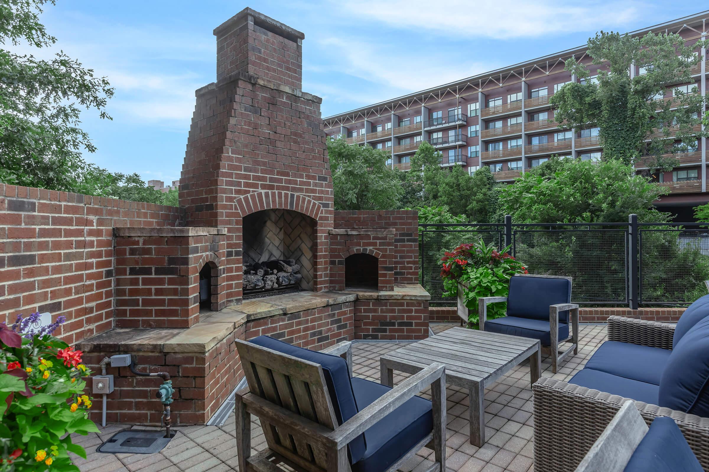 Outdoor patio area featuring a brick fireplace, surrounded by lush greenery and flowering plants. Comfortable seating arrangement with blue cushions and a coffee table. In the background, a modern building can be seen, adding to the inviting atmosphere of the space.