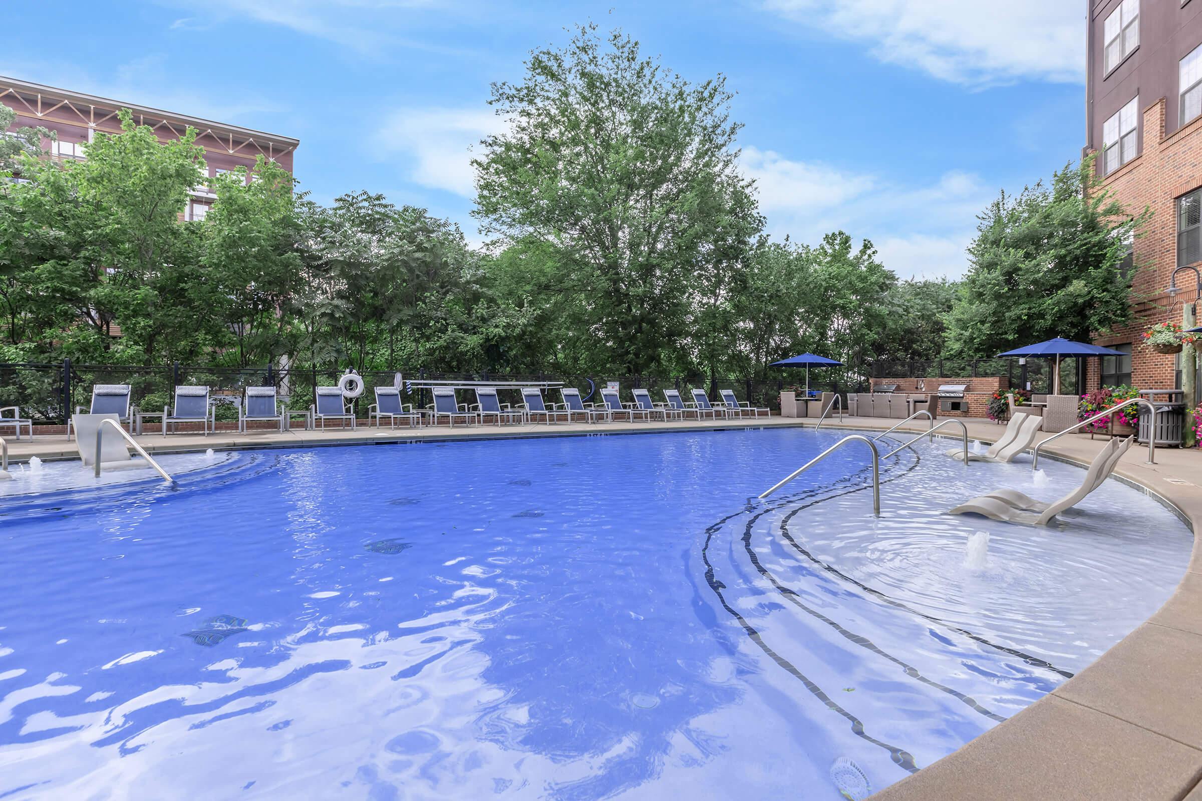 A serene outdoor swimming pool surrounded by lounge chairs and shaded areas with trees in the background. The water is clear and inviting, reflecting the blue sky overhead. The setting suggests a relaxing environment for leisure and recreation.