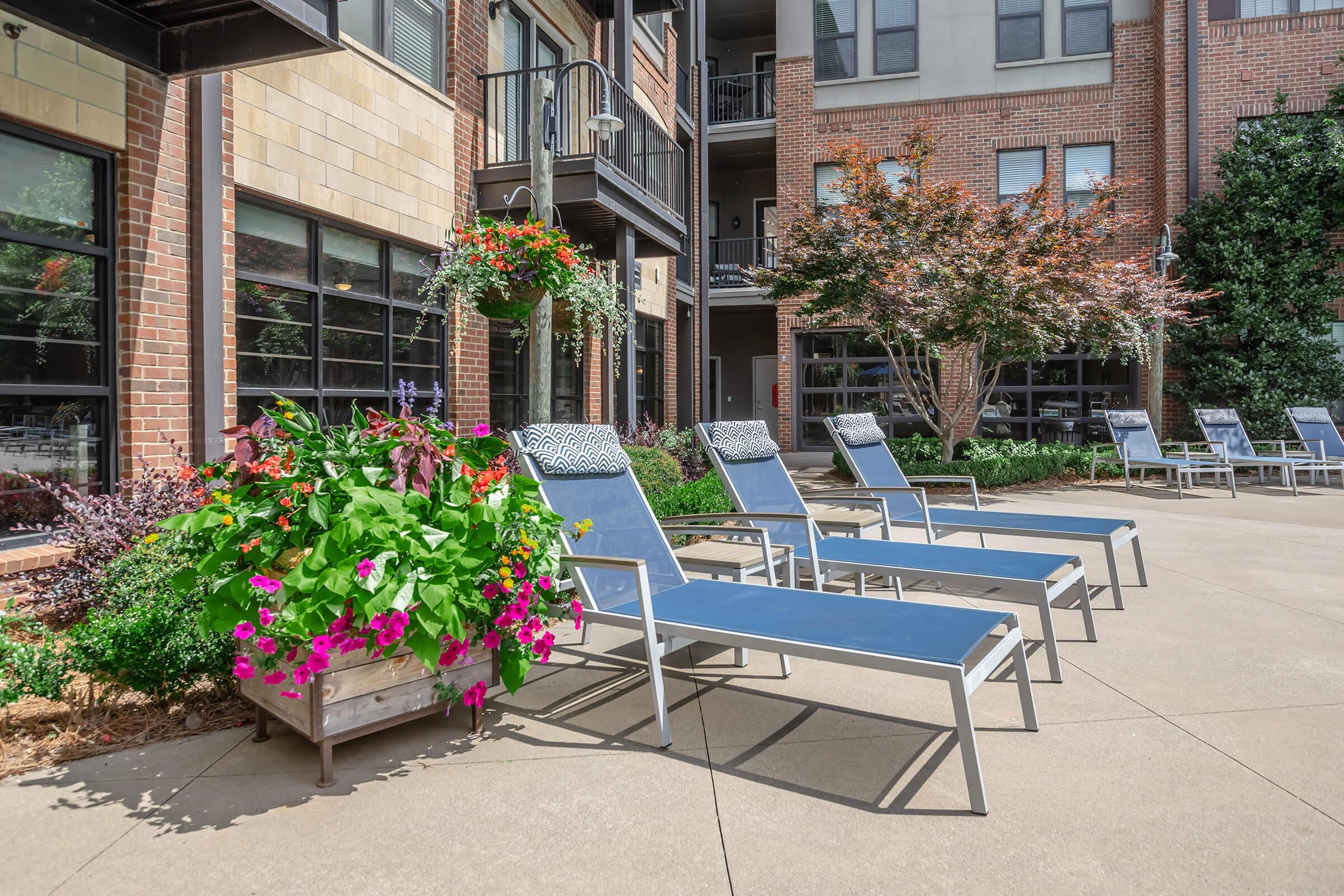 A sunny outdoor area featuring several blue lounge chairs arranged beside vibrant flower beds. The backdrop includes modern apartment buildings with large windows and decorative plants, creating a relaxing atmosphere in a landscaped courtyard.