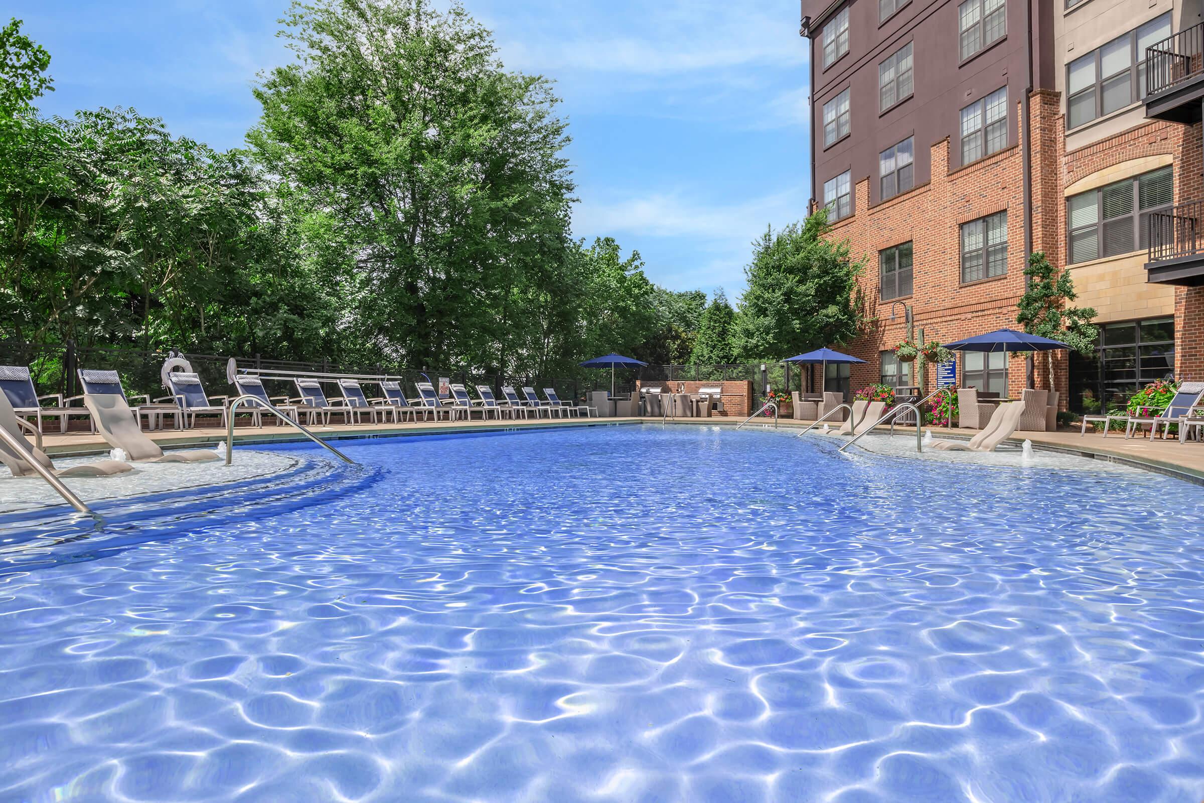 A large swimming pool surrounded by lounge chairs and shaded umbrellas. Lush greenery and trees are visible in the background, alongside a multi-story brick building. The sky is clear and blue, creating a serene atmosphere perfect for relaxation.