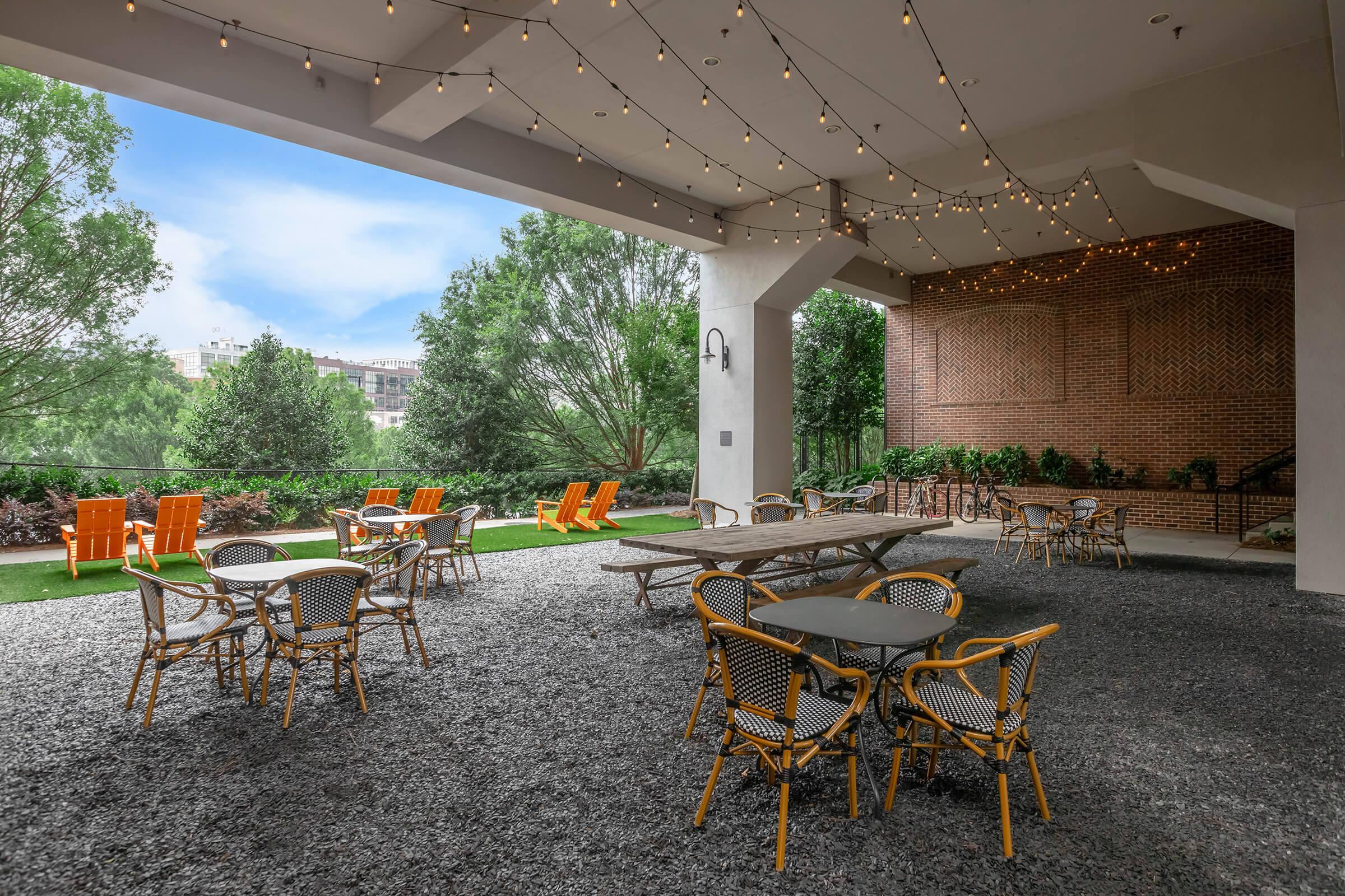 Outdoor patio area featuring a mix of wooden and metal tables with chairs. String lights hang above, creating a cozy atmosphere. The space is surrounded by greenery, with several orange lounge chairs visible in the background. The ground is covered in gravel, adding to the rustic vibe.