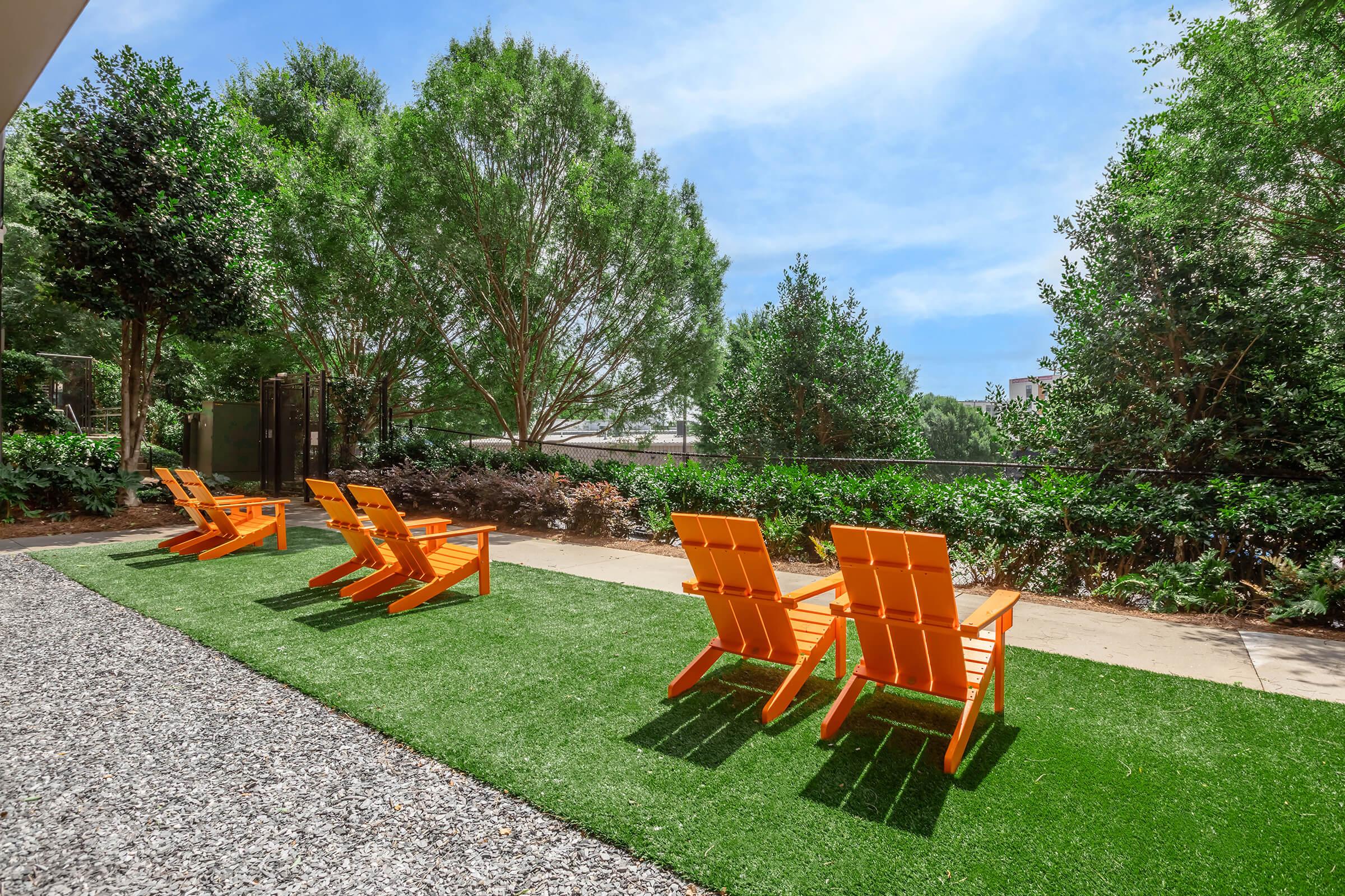 Four bright orange lounge chairs on a green, grassy area surrounded by trees and shrubs, under a clear blue sky. The chairs are arranged on a path made of gravel, creating a relaxing outdoor space.