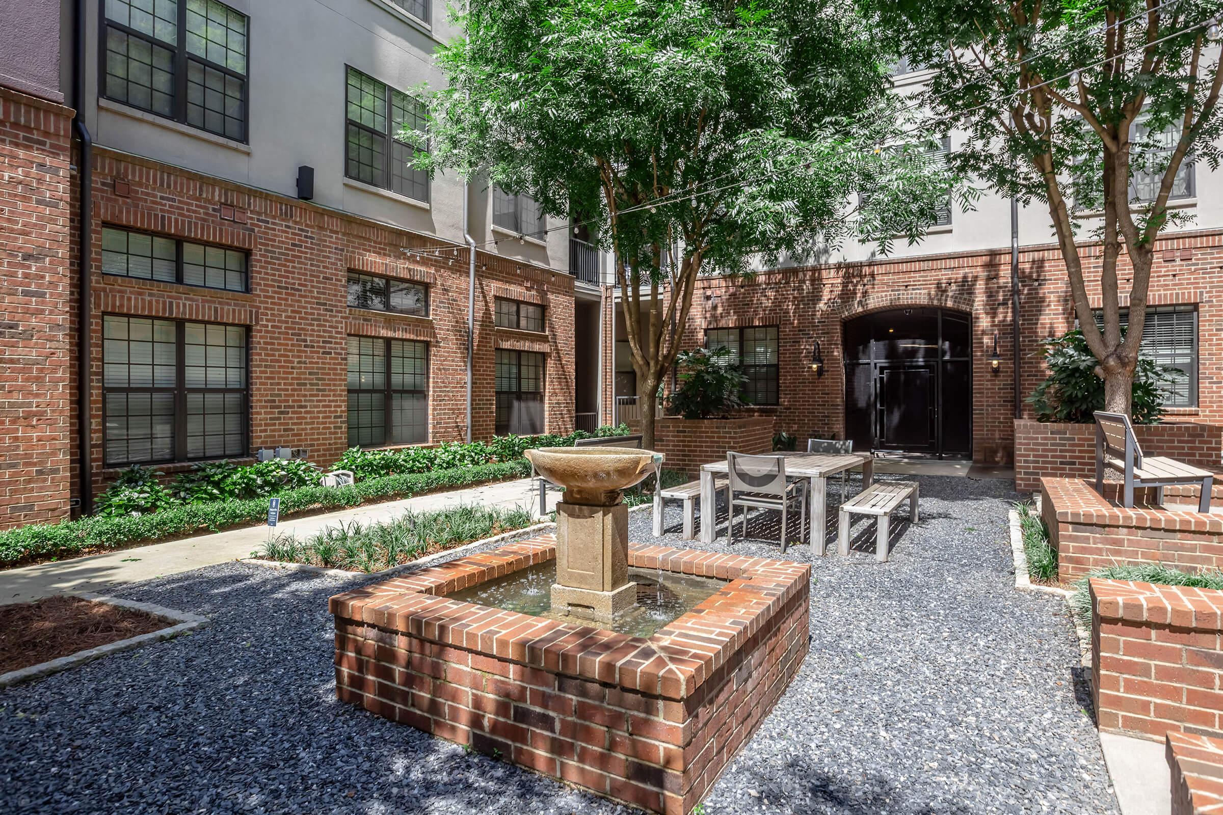 A serene courtyard featuring a small water fountain at the center, surrounded by brick seating and gravel pathways. Lush greenery and trees provide a tranquil atmosphere, with brick buildings in the background. The design creates a peaceful outdoor space for relaxation and gatherings.