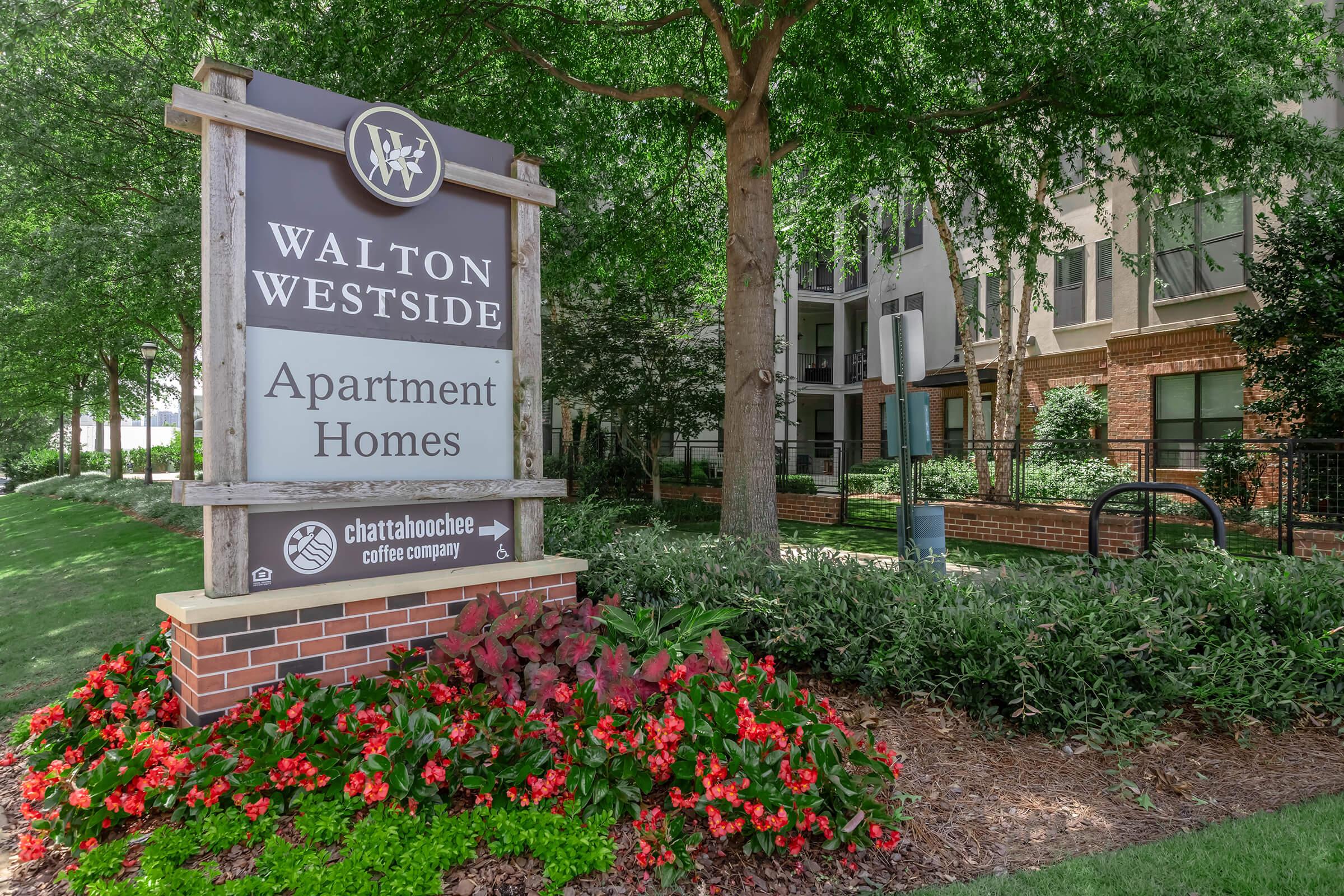 Sign for Walton Westside Apartment Homes, featuring the name and logo prominently. Surrounding the sign are well-maintained green shrubs and colorful flower beds. The background includes residential buildings under a clear sky.
