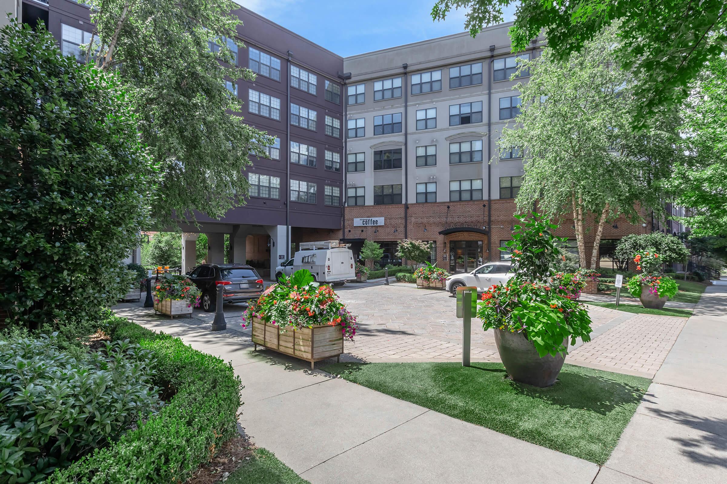 A well-maintained urban apartment complex featuring a landscaped entrance with flower planters, greenery, and parked cars. The building showcases a mix of modern architecture and brick, with large windows letting in natural light. Clear blue skies add to the inviting atmosphere of the setting.