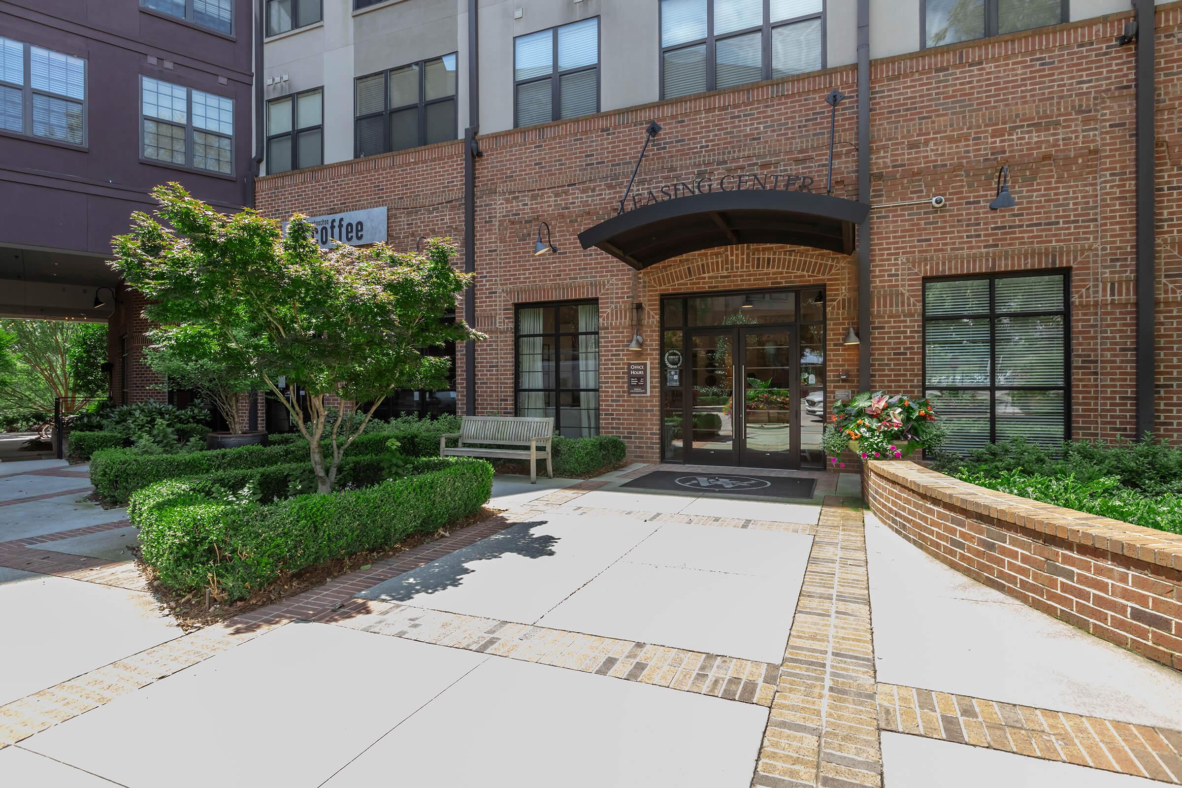 A brick building entrance with large windows and a dark awning. The area is landscaped with greenery, including bushes and flower pots. A wooden bench is positioned nearby on a paved walkway leading to the entrance, which has a sign above it.