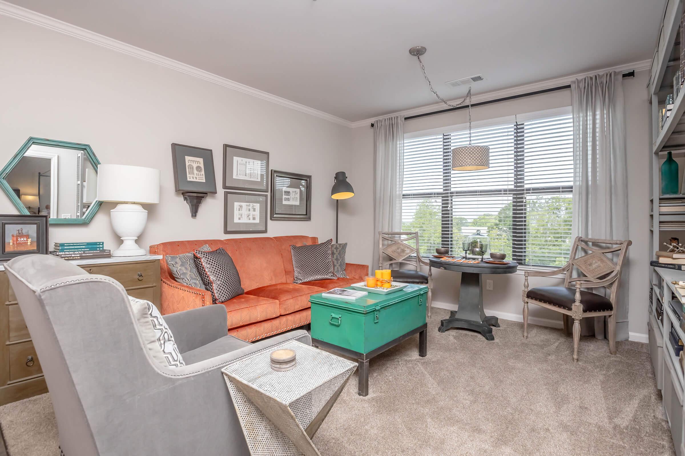 A cozy living room featuring a bright orange sofa, a teal coffee table, and a mix of decorative items on the walls. The space includes two stylish chairs, a lamp, and a large window with blinds letting in natural light, creating a warm and inviting atmosphere.