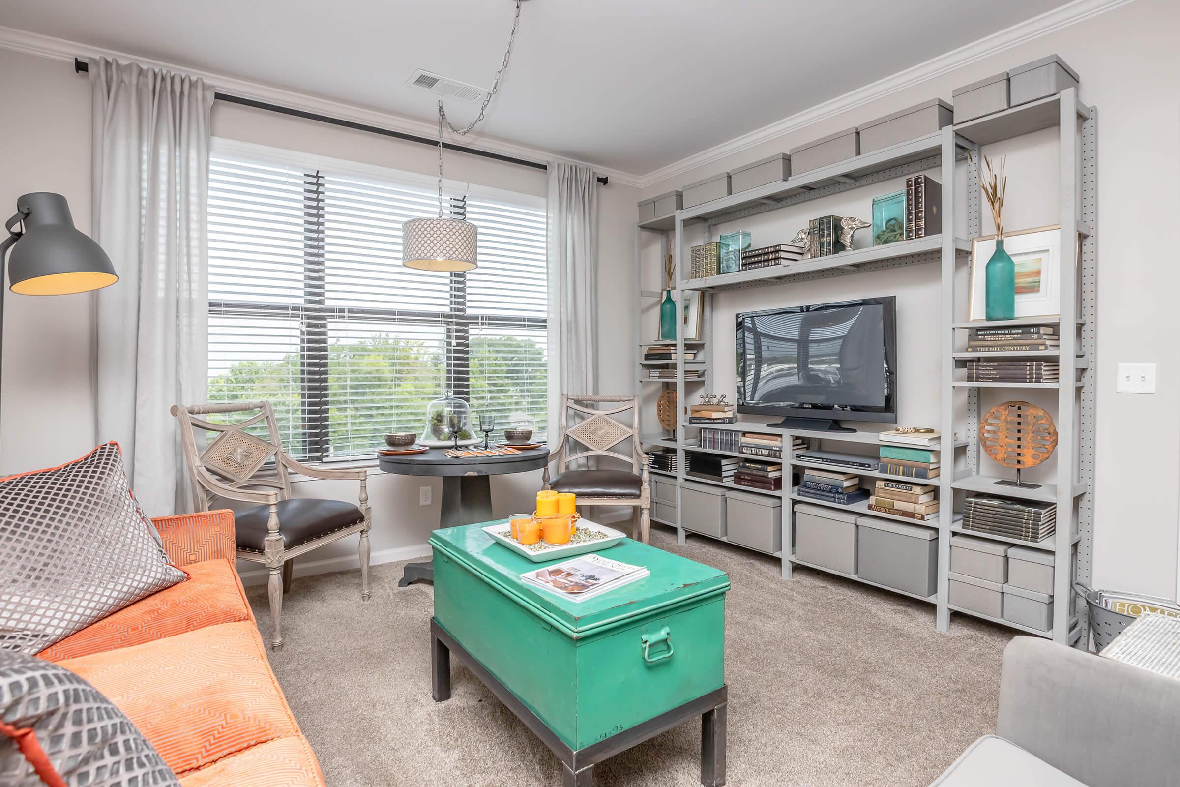 A stylish living room featuring a bright orange sofa, a green storage chest, and a wall-mounted television. The room has large windows with sheer curtains, a decorative bookshelf filled with books and decor items, and a coffee table with a stack of magazines and a tray of orange drinks.