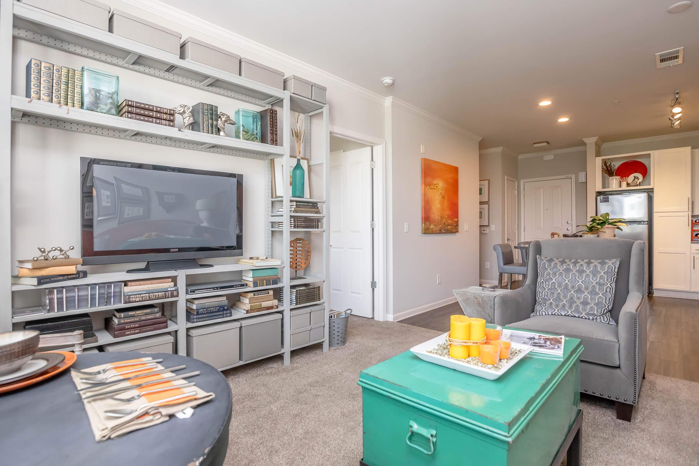 A cozy living room featuring a gray armchair, a turquoise coffee table, and a modern TV stand filled with books and decor. The space has soft carpet, light-colored walls, and a dining area in the background with kitchen access. Decorative items and a warm color scheme create an inviting atmosphere.