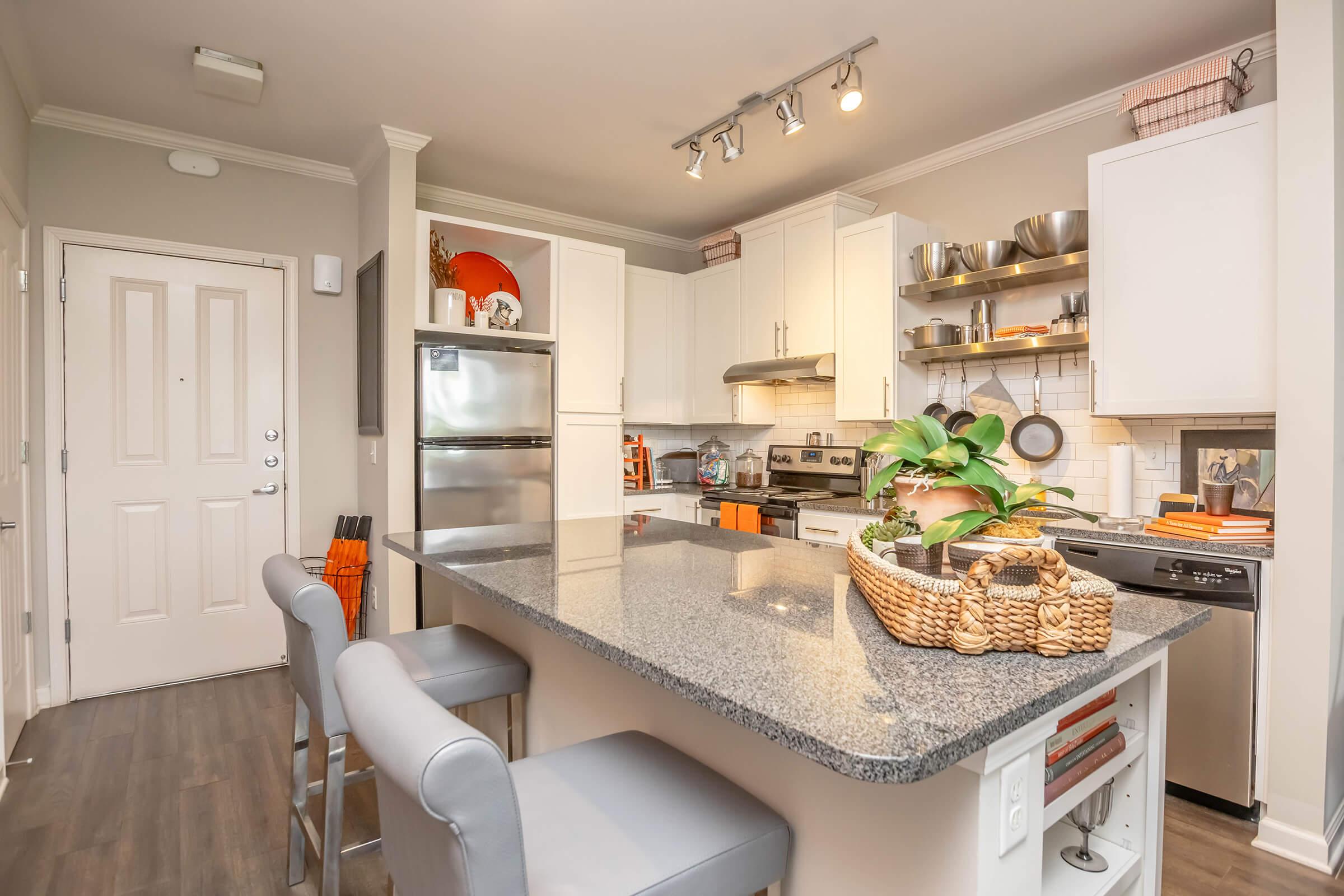 Modern kitchen featuring white cabinetry, stainless steel appliances, and a large granite island with seating. The decor includes a woven basket with plants, open shelving showcasing kitchenware, and a welcoming entrance with a door and light fixtures.