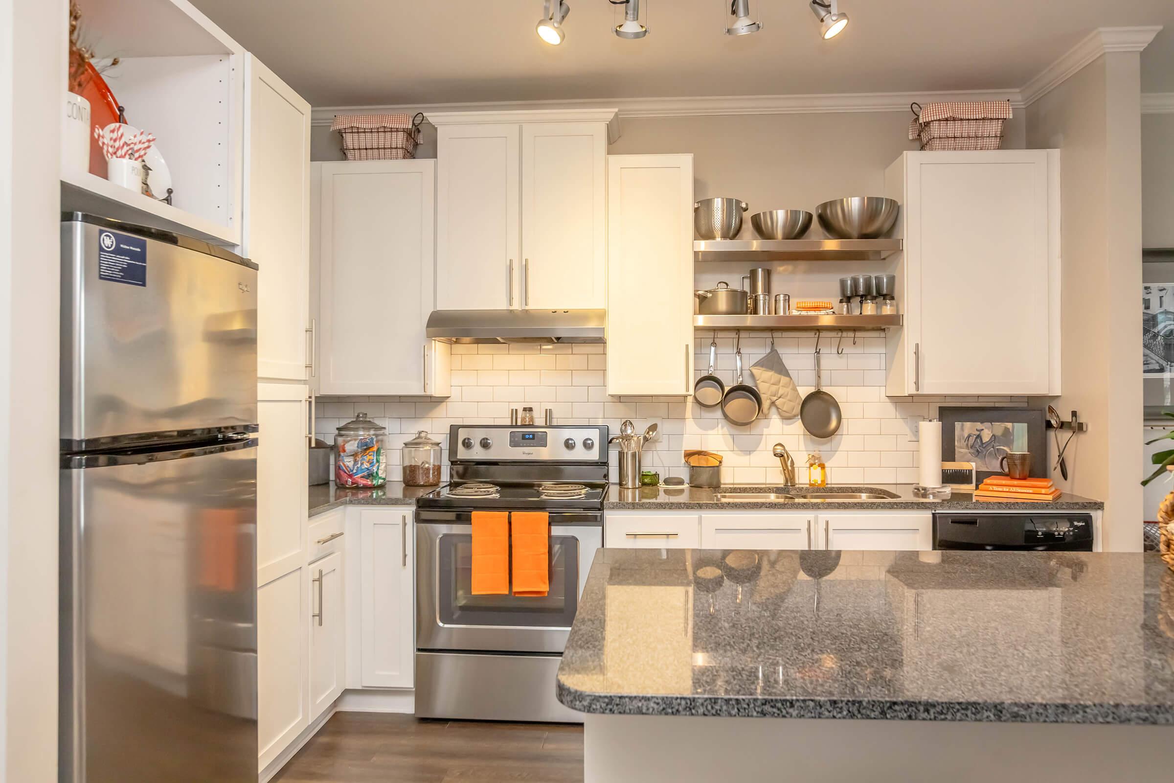 A modern kitchen featuring white cabinets, stainless steel appliances, and a granite countertop. The kitchen includes an oven, stove, and sink, with various cooking utensils and bowls displayed on open shelves. An orange towel is hanging on the oven handle, adding a pop of color to the clean, organized space.