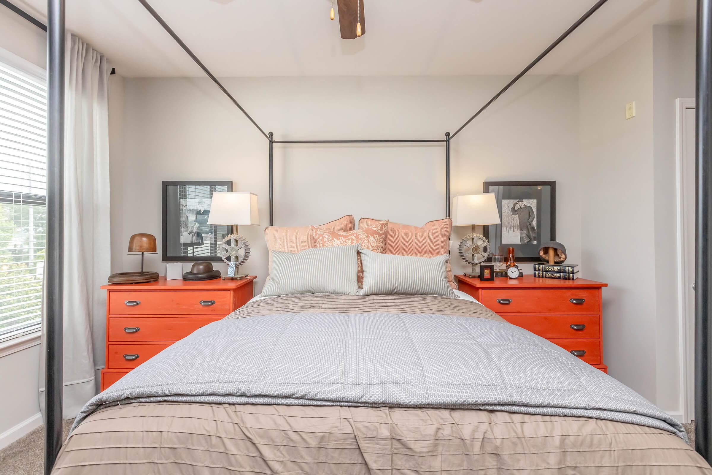 A cozy bedroom featuring a four-poster bed with neutral bedding. On either side of the bed are orange dressers with decorative items and lamps. The room has light gray walls, large windows with sheer curtains, and framed artwork on the walls, creating a warm and inviting atmosphere.