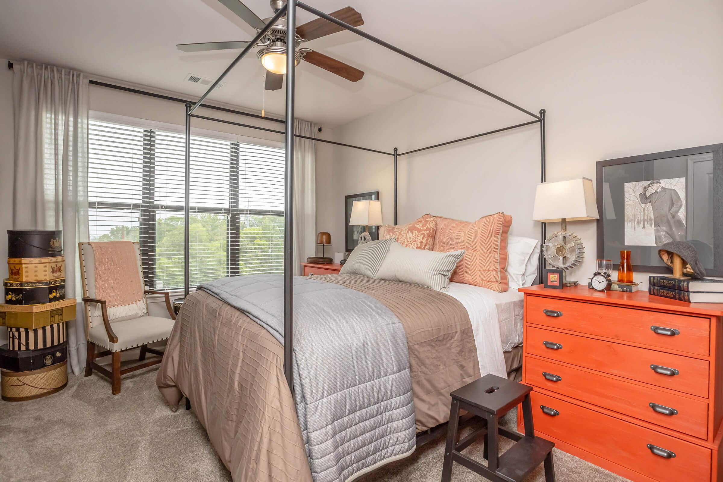 A cozy bedroom featuring a four-poster bed with neutral bedding, an orange accent dresser, and a comfortable armchair. The room is well-lit by natural light from large windows, with curtains that add a soft touch. Decorative elements include a table lamp, artwork, and various small decor items.