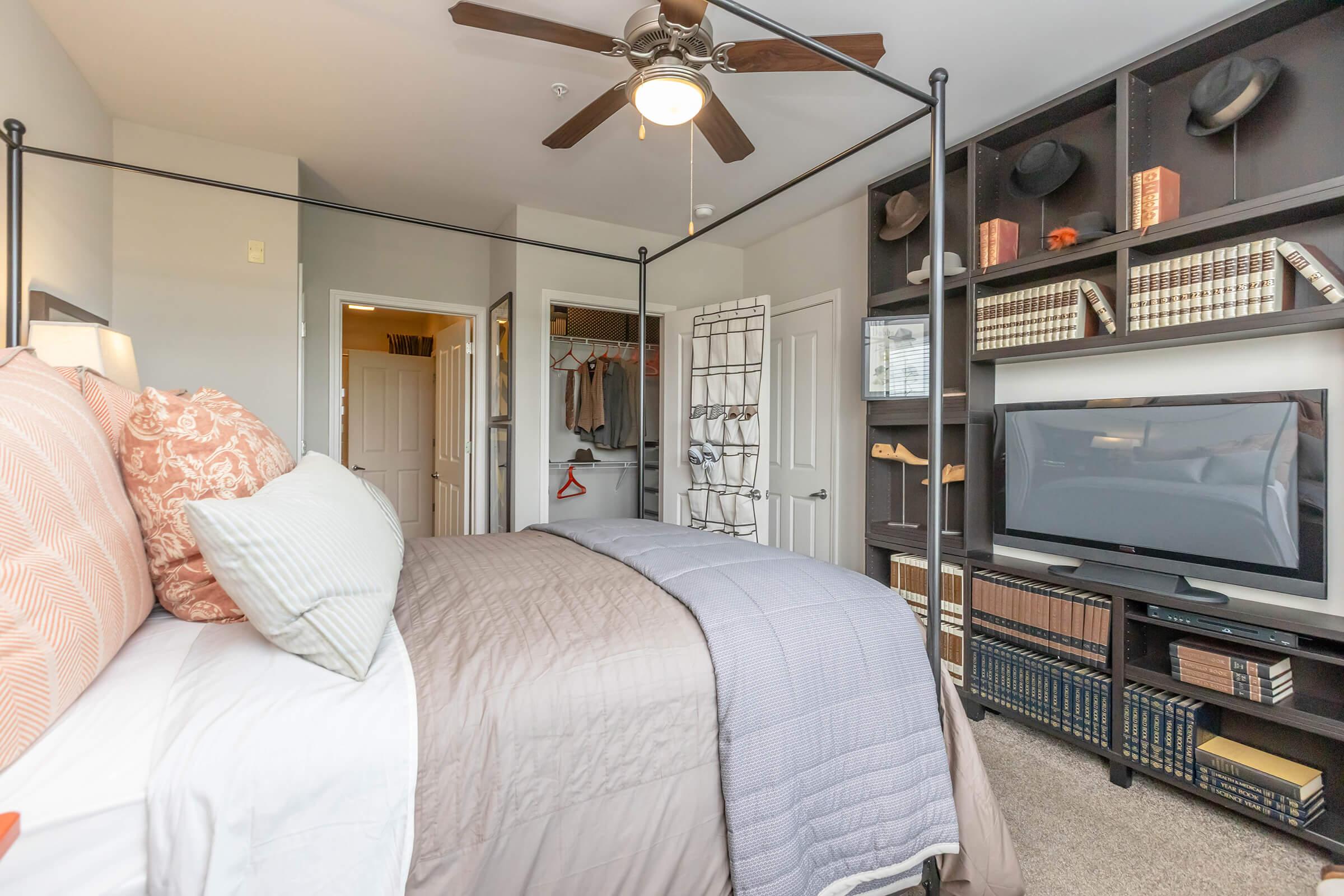 A cozy bedroom featuring a four-poster bed with decorative pillows, a ceiling fan, and light gray bedding. There's a closet with hanging clothes visible, a shelf with books and hats, and a television mounted on the wall. The room has a warm and inviting atmosphere.