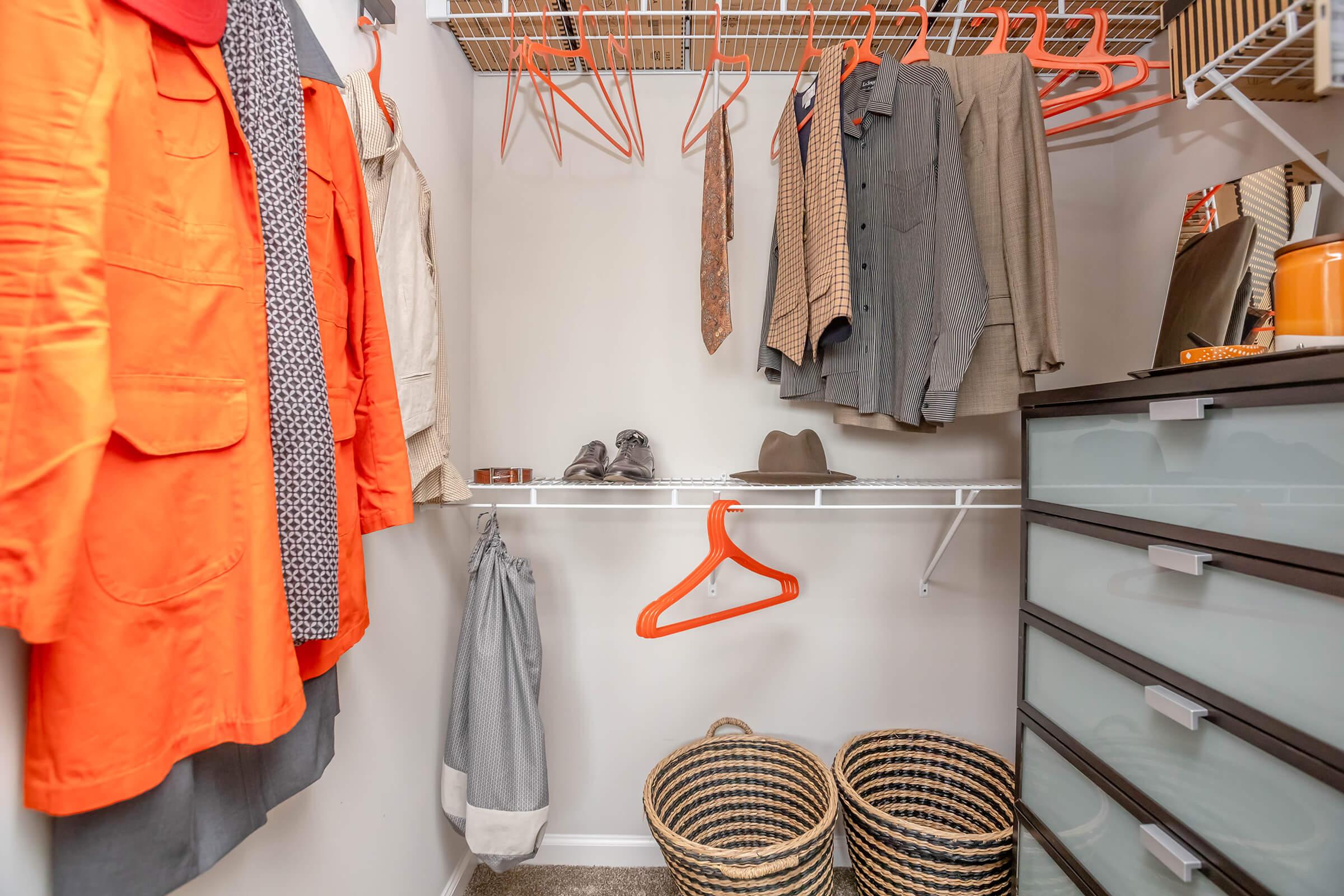 A neatly organized closet featuring hanging clothing items, including an orange jacket, shirts, and ties. On the shelf, there are gray shoes and a decorative item. Below, two woven baskets are stored, and a gray garment bag is hanging on a white rack, contributing to a tidy appearance.