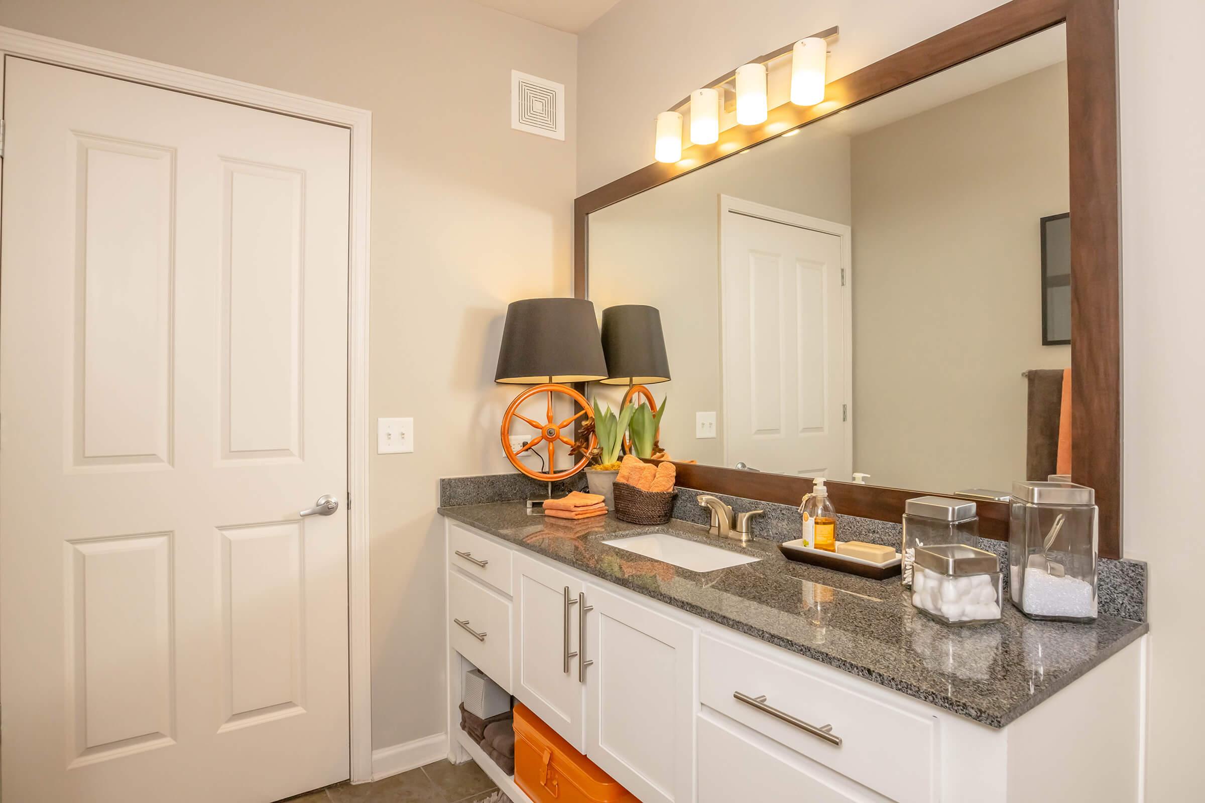 A modern bathroom featuring a gray granite countertop with a large mirror above it. There are stylish fixtures, a lamp with a decorative base, a potted plant, and neatly arranged towels. An adjacent door is visible, and there are storage areas with orange accents. The walls are painted in a neutral tone, creating a bright atmosphere.