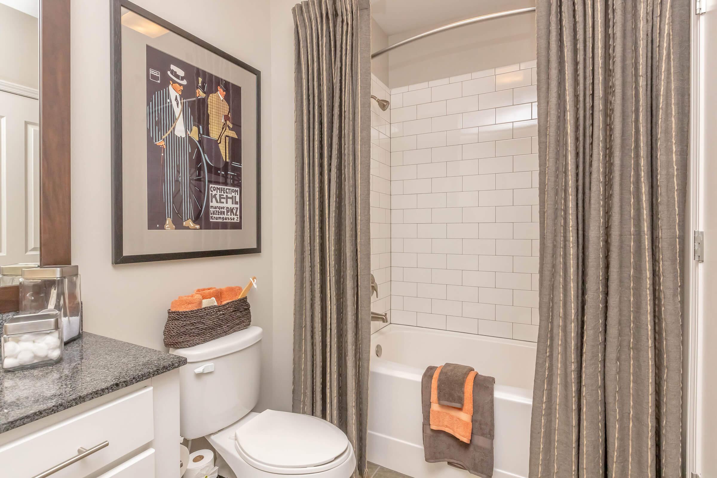 A modern bathroom featuring a bathtub with a shower, surrounded by beige and white tiles. The space includes a dark countertop with storage containers and decorative items. A framed vintage-style poster hangs on the wall, and there are gray and orange towels displayed. The curtains are gray with subtle vertical stripes.