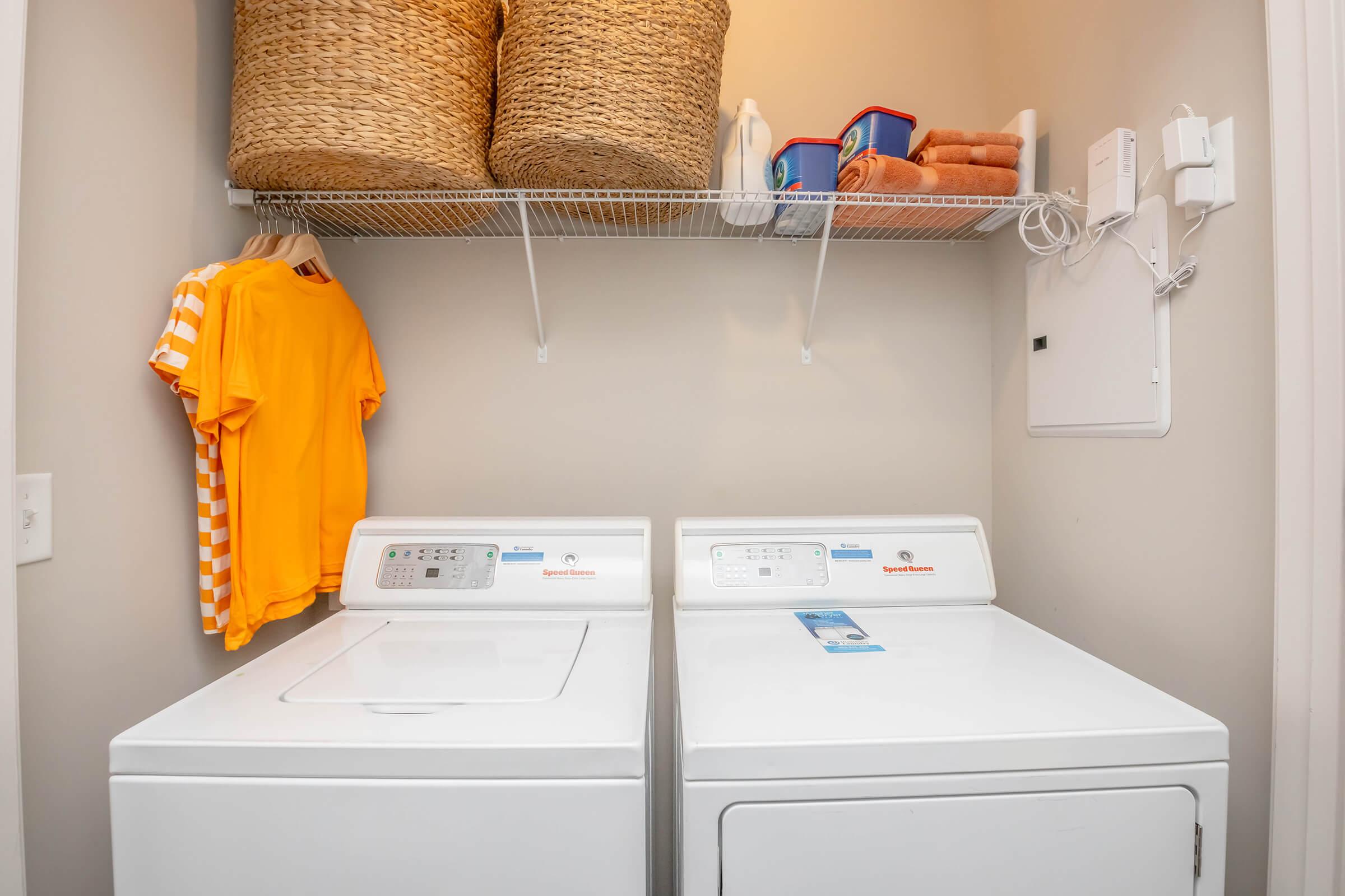 A laundry room featuring a washing machine and dryer side by side. Above them, a wire shelf holds two woven baskets and cleaning supplies. A bright orange shirt hangs on the wall next to the machines, and neatly stacked towels are visible on the shelf. The walls are painted a soft neutral color.