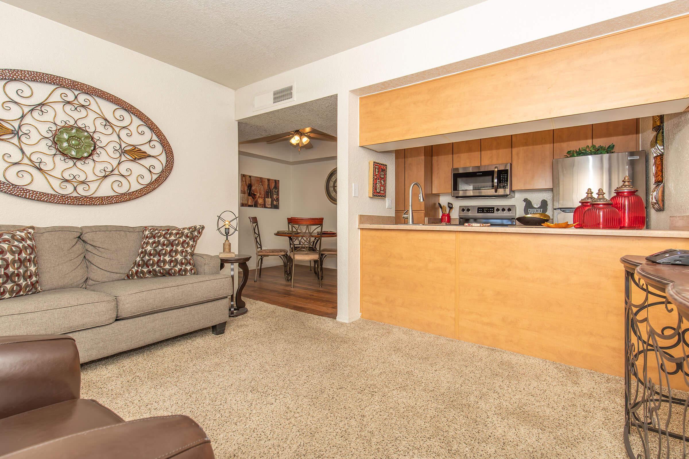 Cozy living area featuring a beige sofa with patterned cushions, a small dining area in the background, and an open kitchen with wooden cabinetry and stainless steel appliances. Decorative wall art and a plush carpet enhance the welcoming atmosphere of the space.