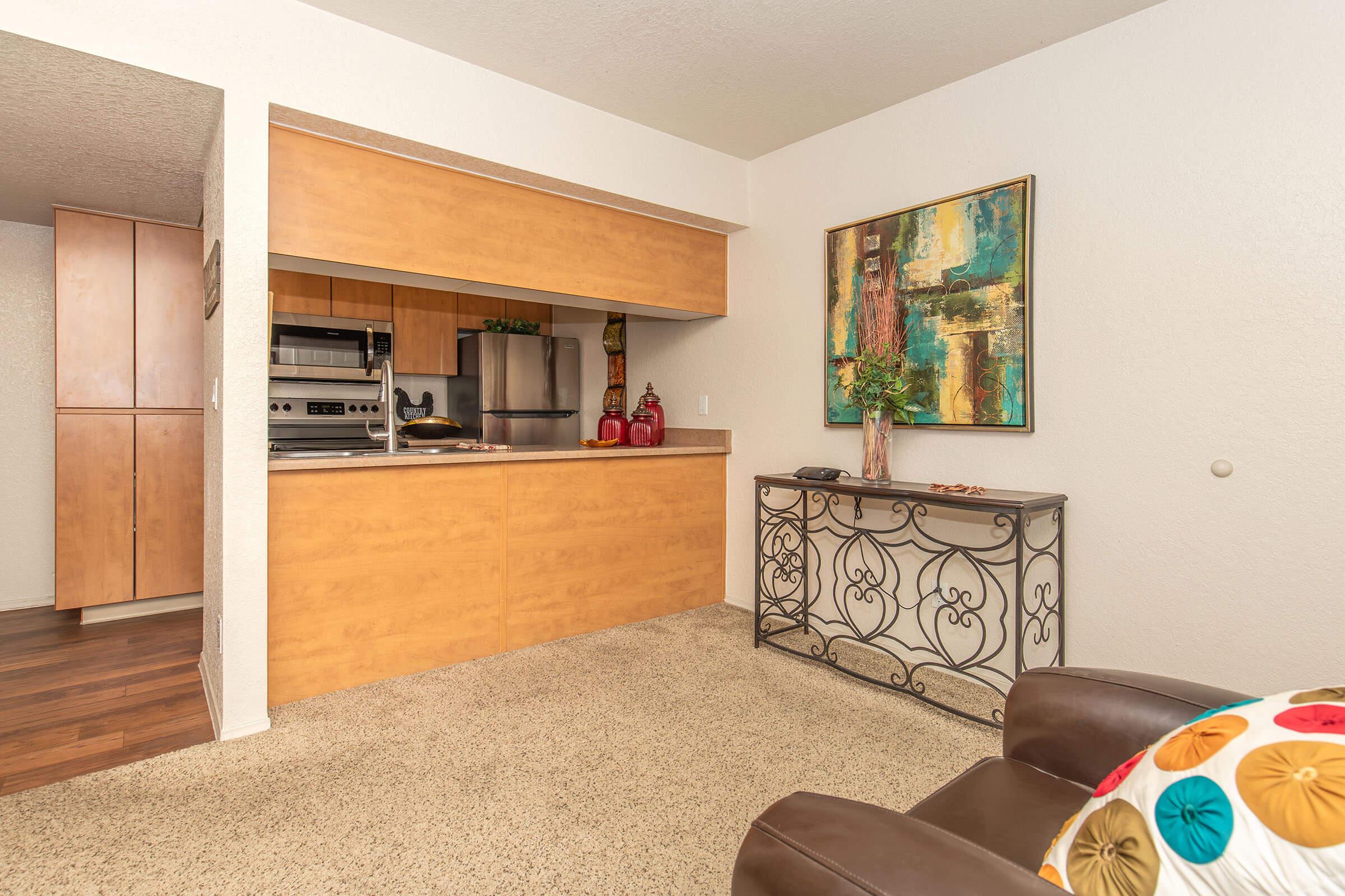 An interior view of a cozy living space featuring a small kitchen area with wooden cabinetry, stainless steel appliances, and decorative items. A stylish console table is positioned against the wall, and there is a comfortable brown chair with a colorful pillow in the foreground, creating a welcoming atmosphere.