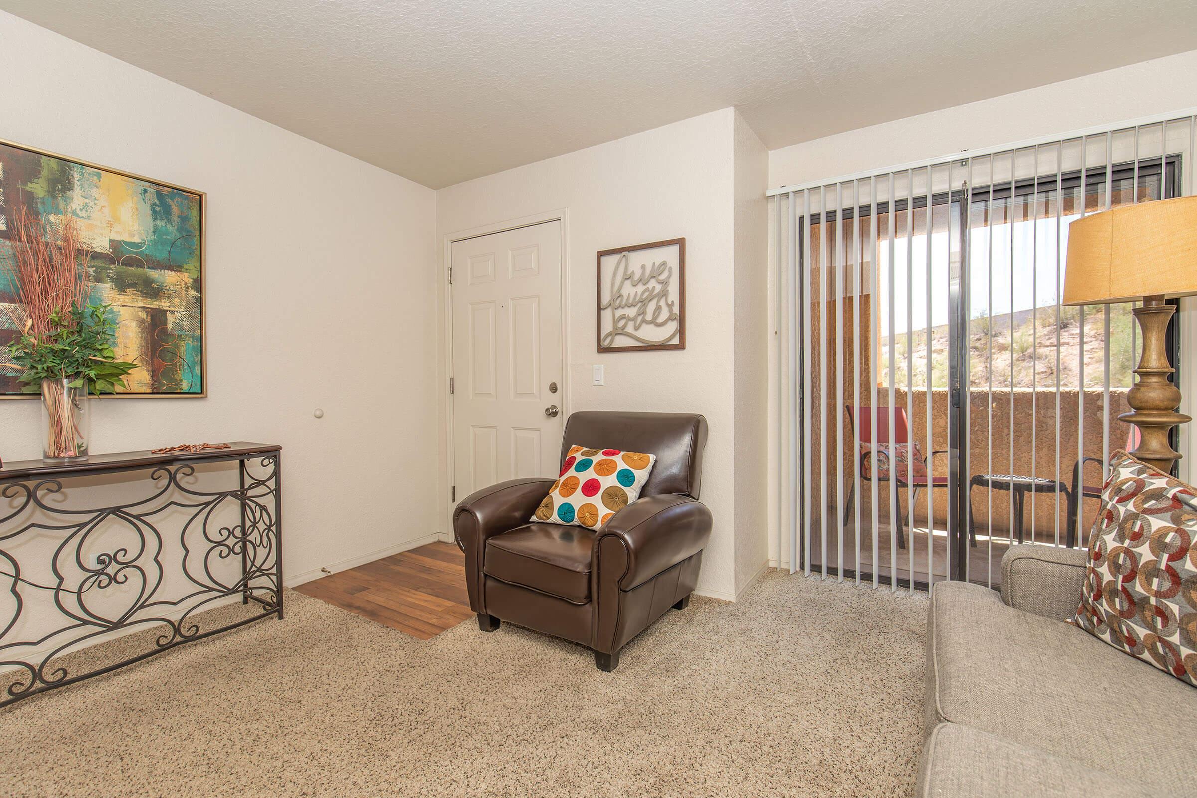 A cozy living room featuring a brown leather chair with colorful cushions, a decorative console table with artwork above it, and a beige sofa. The room has light-colored walls and a door leading to a balcony, with sheer vertical blinds allowing natural light to enter. The flooring is carpeted.