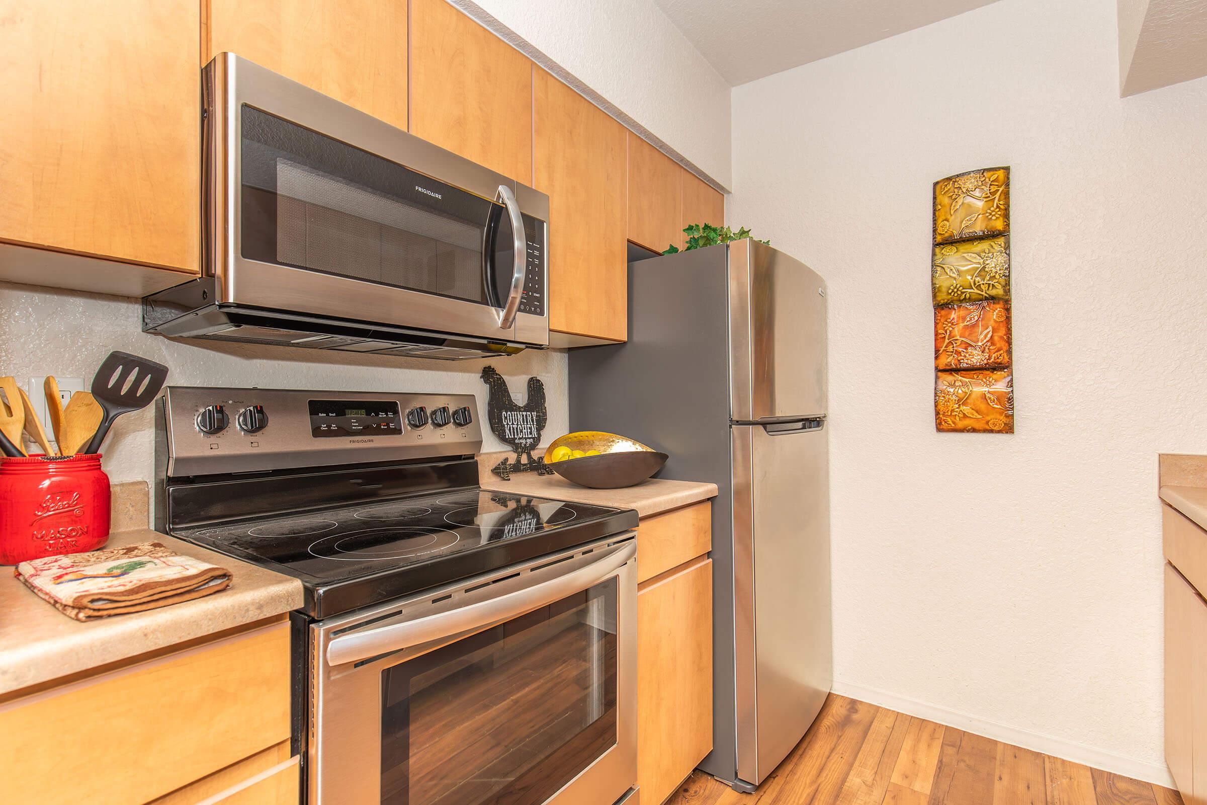 A modern kitchen featuring stainless steel appliances, including a microwave and oven. The wood cabinetry complements the light-colored walls. A bowl of fruit sits on the countertop, and a colorful wall art piece adds a touch of decor. Warm hardwood flooring completes the inviting atmosphere.