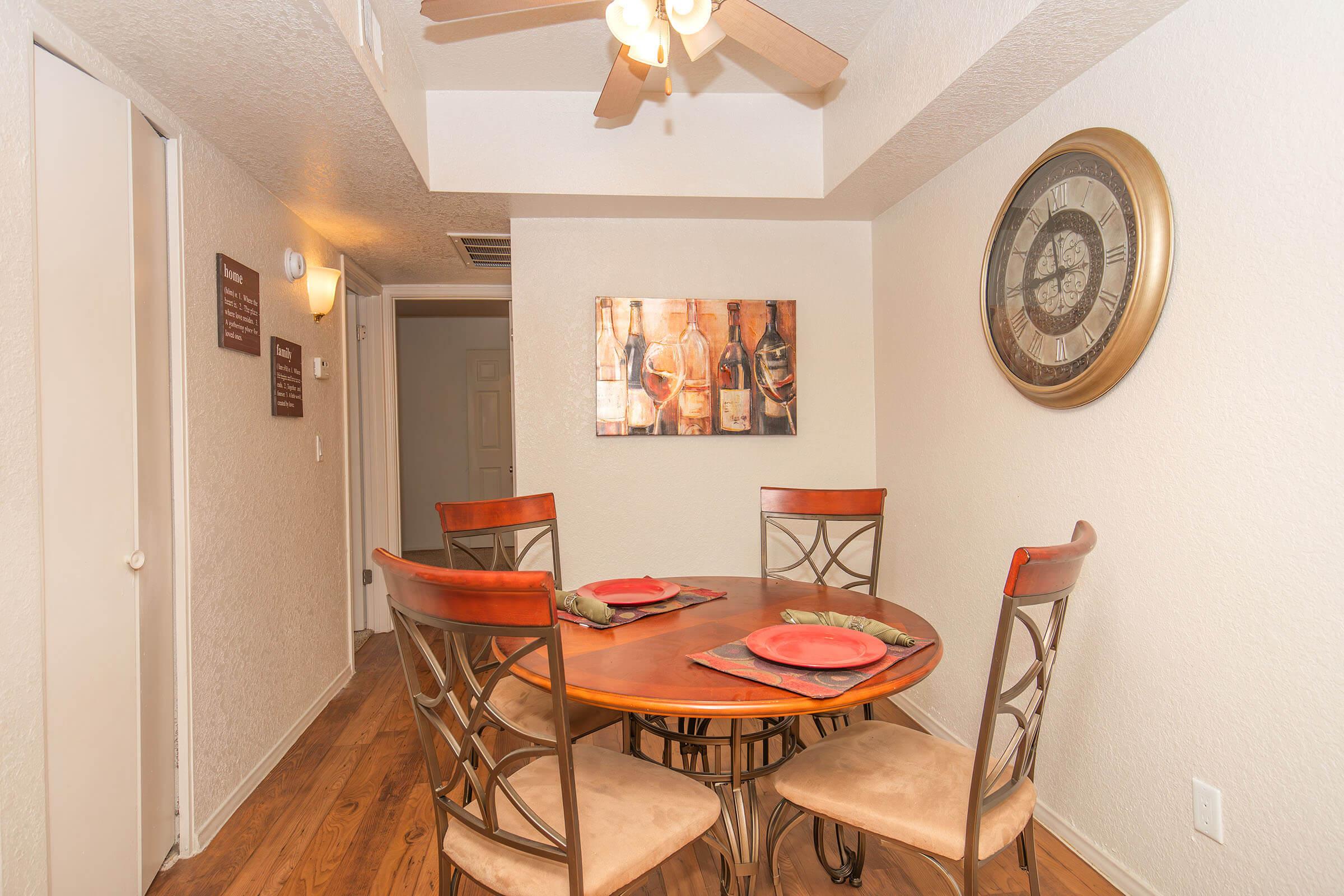 A dining area featuring a round wooden table with four upholstered chairs, set with plates and napkins. The walls are decorated with a large clock and framed artwork of wine bottles. Soft lighting comes from a ceiling fan. The floor is wooden, and there are light-colored walls and a door in the background.