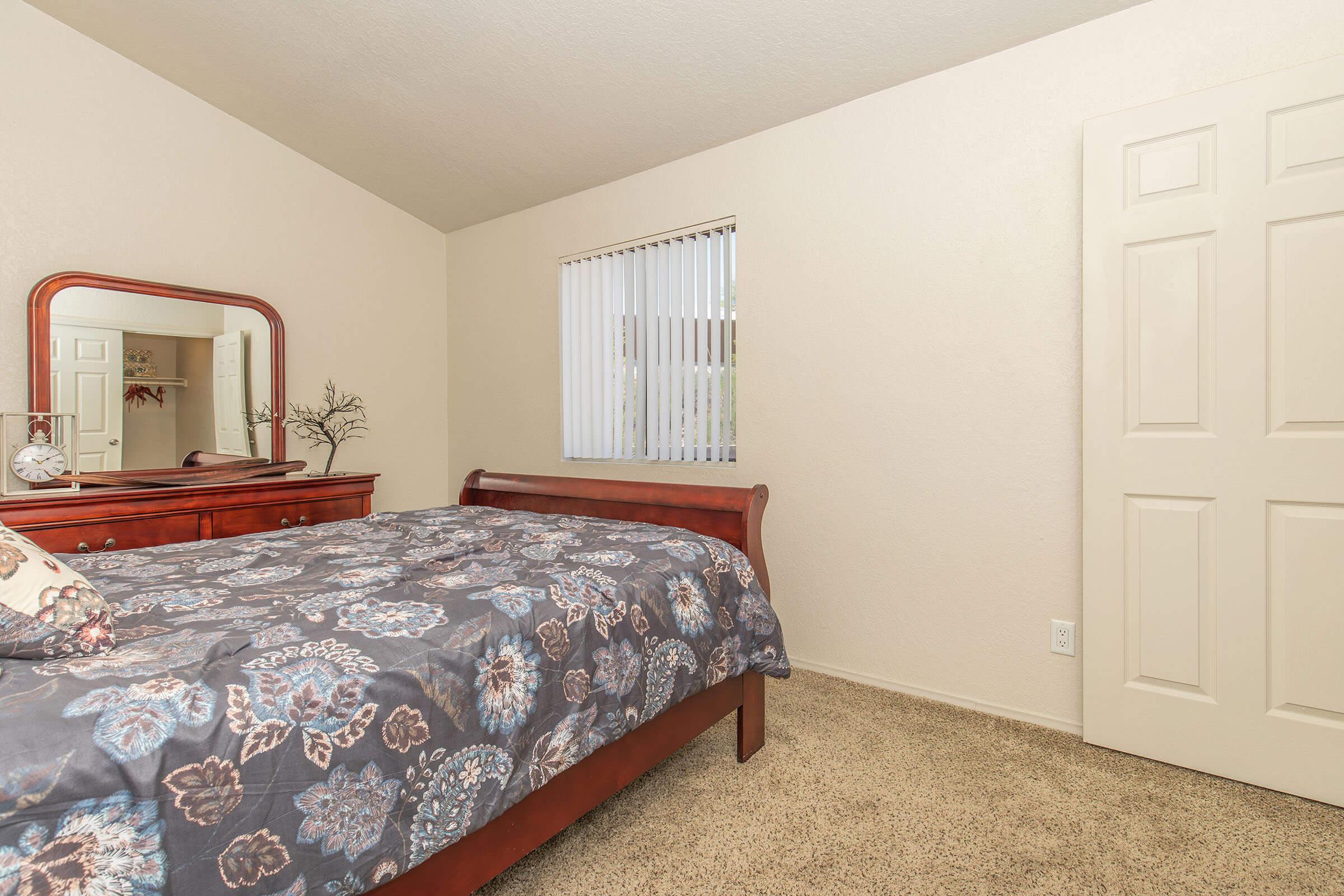A cozy bedroom featuring a wooden sleigh bed with a floral patterned comforter, a matching dresser with a mirror, and a window with vertical blinds. The walls are painted in a light neutral color, and the carpet is beige, adding warmth to the space. A closed door is visible on one side.