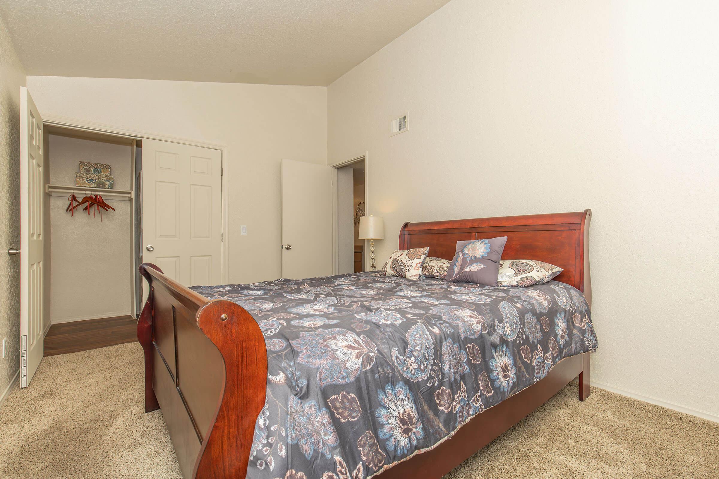A cozy bedroom featuring a wooden sleigh bed with a floral patterned comforter. The walls are painted a light color, and there is a lamp beside the bed. A closet is visible in the background, with a door partially open, revealing additional storage space. The carpet is soft and beige, creating a warm ambiance.