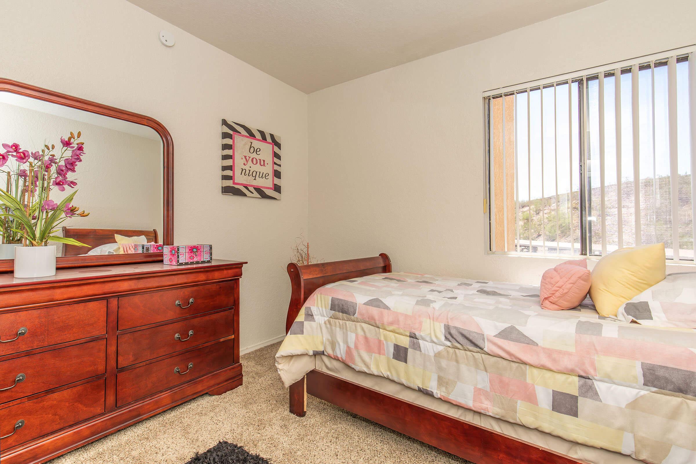 A cozy bedroom featuring a wooden bed with a multicolored quilt, a matching wooden dresser, and a large mirror. A vase with flowers sits on the dresser, while a framed motivational poster hangs on the wall. Sunlight filters through vertical blinds, illuminating the space.