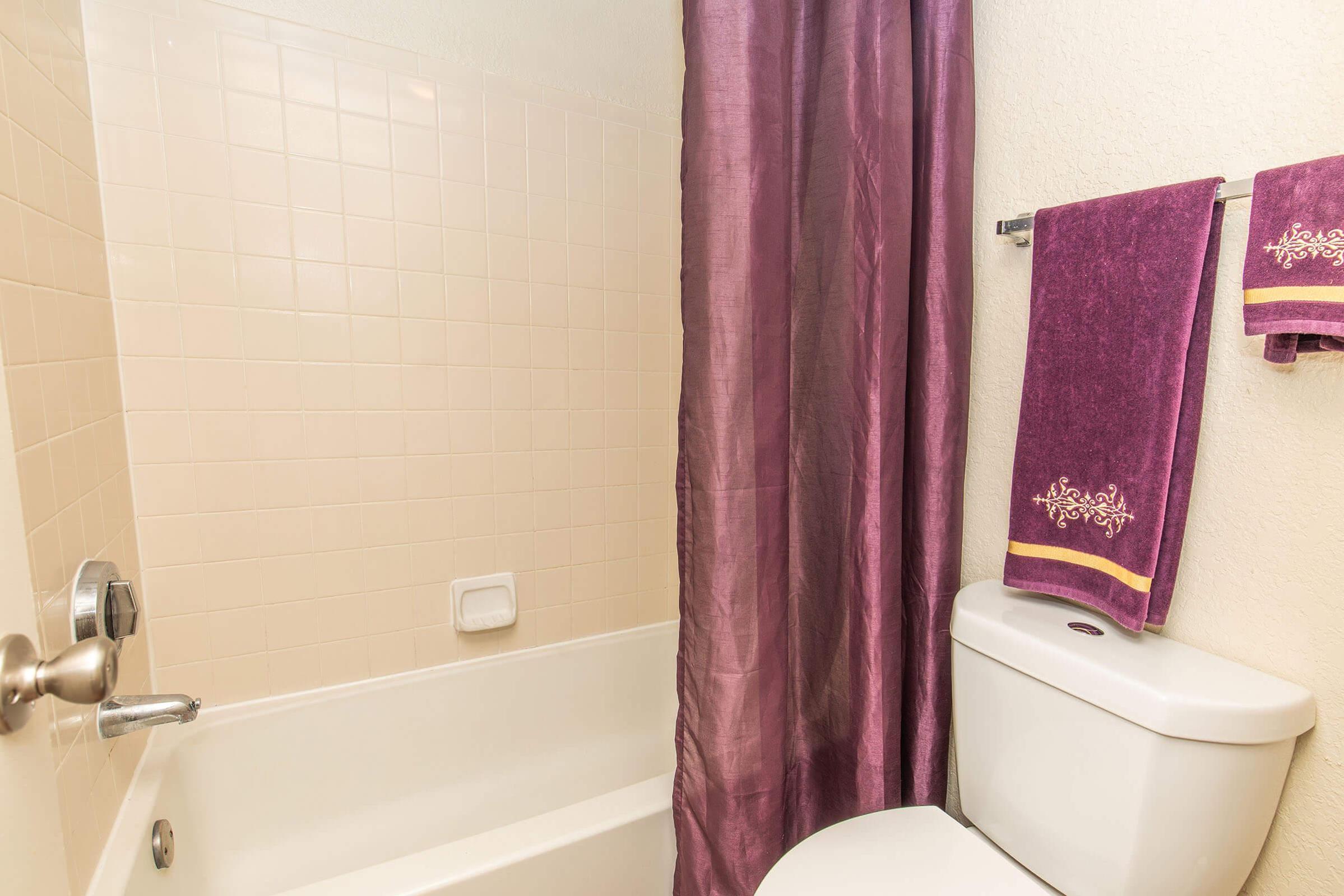A bathroom featuring a white bathtub with a purple shower curtain. There are two purple towels hanging on a rack beside the bathtub, complementing the curtain. A white toilet is situated next to the bathtub, with neutral-colored tiles on the wall. The overall design is clean and well-organized.