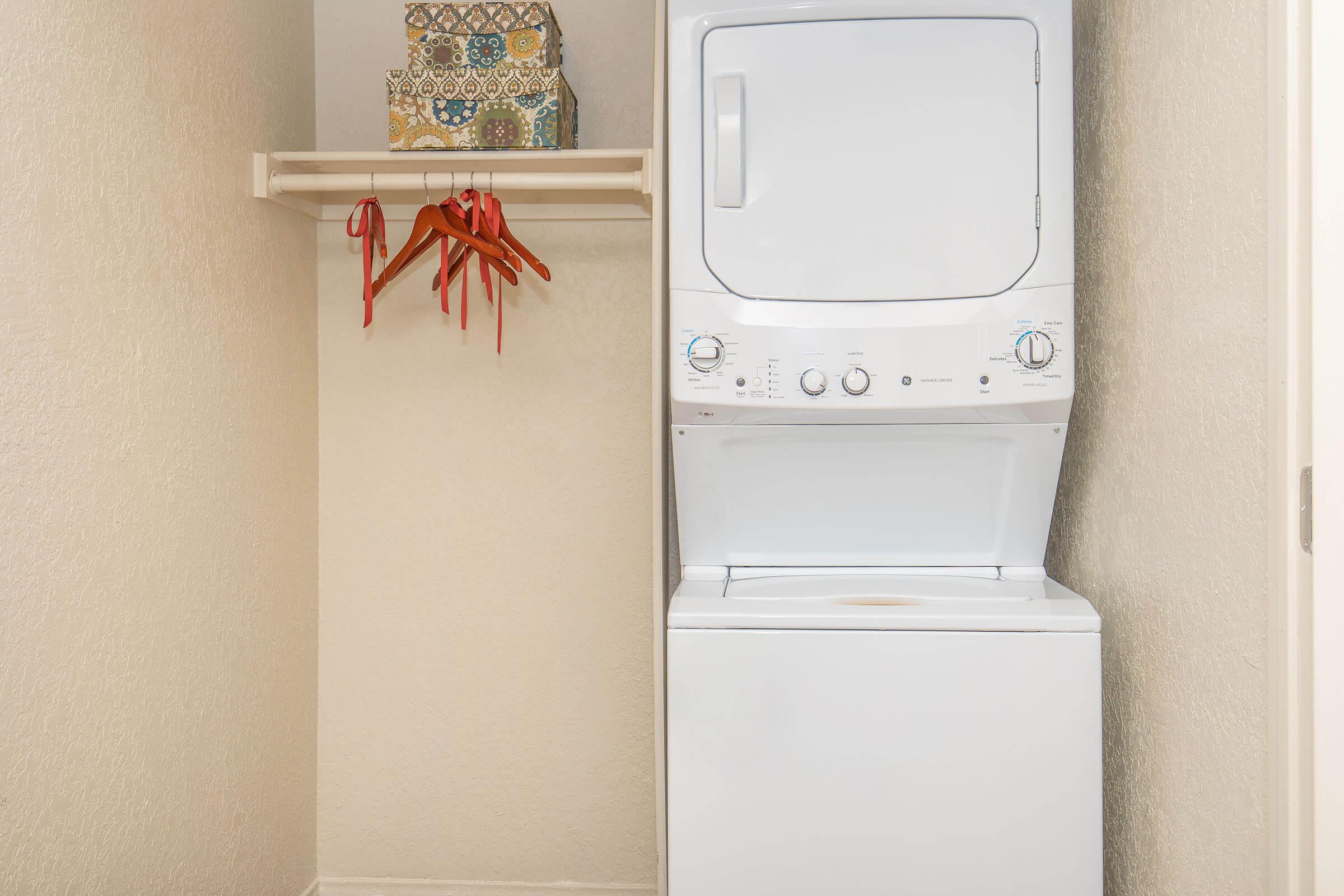 A compact laundry area featuring a stacked washer and dryer unit. Above the appliances, there is a shelf with a decorative box. To the side, a rack holds three red items or accessories, complementing the neutral-toned walls.