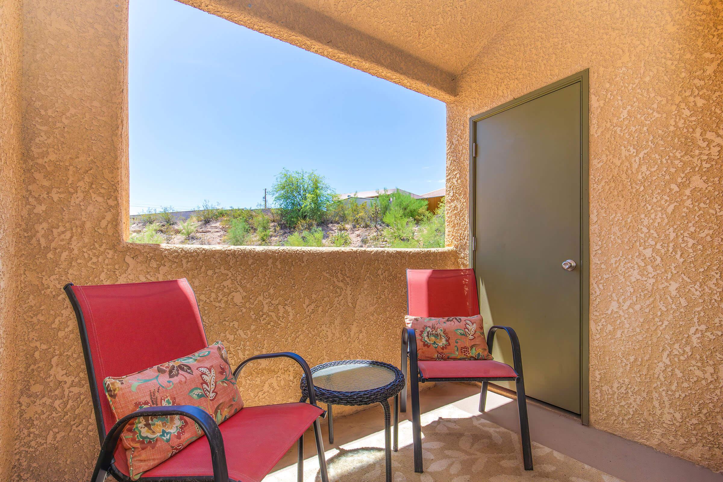 A cozy outdoor patio featuring two red chairs with decorative cushions and a small round table. The patio has a textured beige wall and a large rectangular window that opens to a view of greenery and a blue sky.