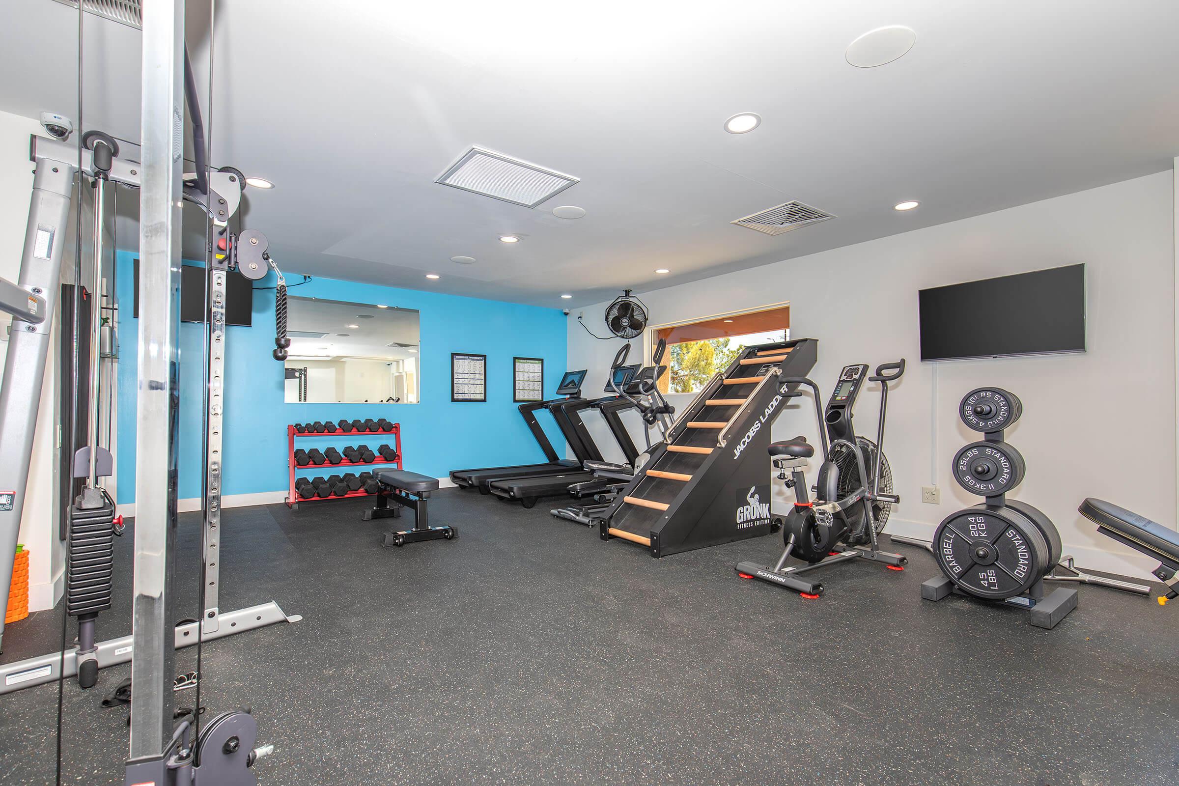 A modern gym interior featuring various exercise equipment, including treadmills, a stair climber, a weight bench, free weights on racks, a stationary bike, and a large wall-mounted TV. The flooring is black rubber, and there's a blue accent wall in the background.