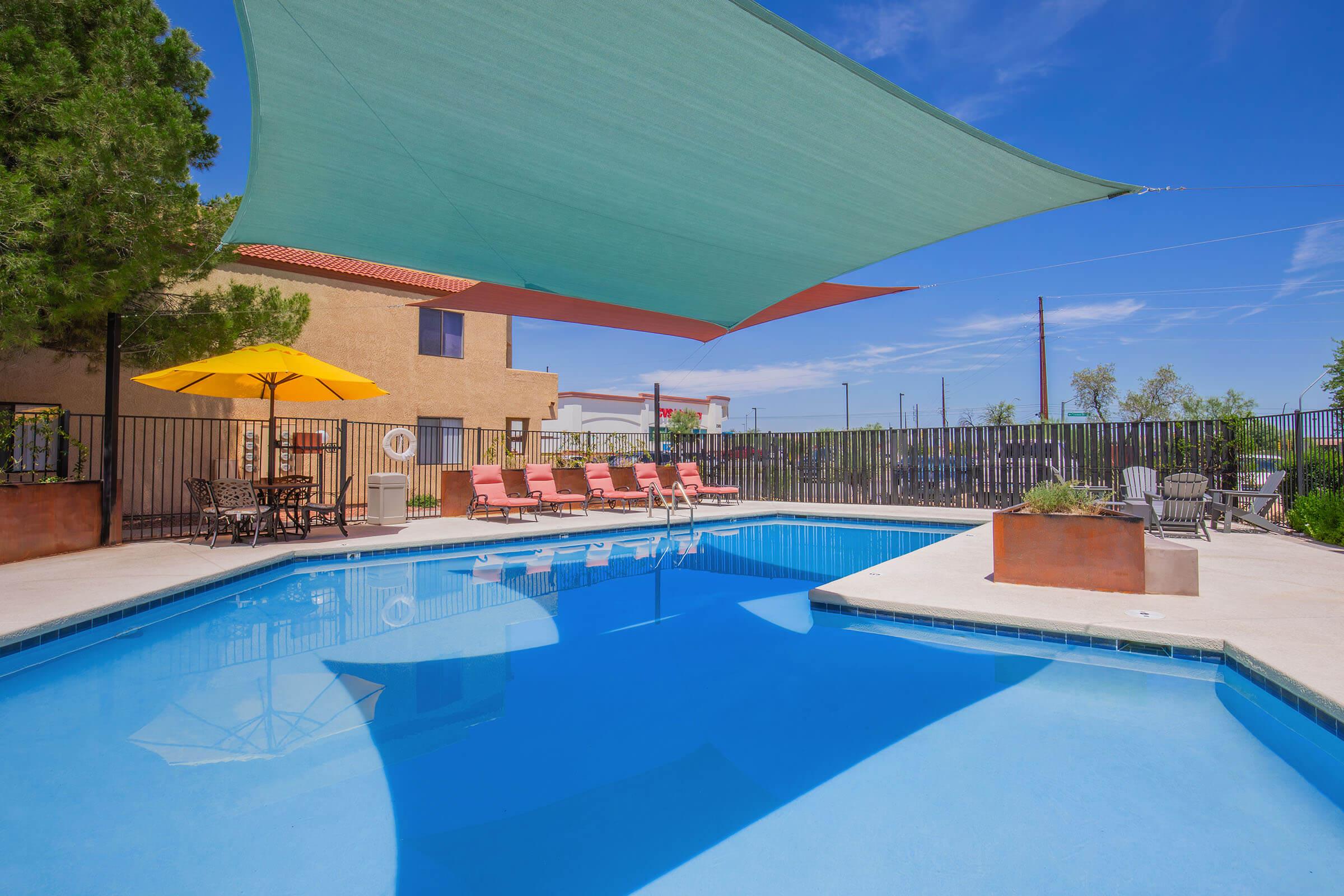 A bright outdoor pool area featuring clear blue water, surrounded by lounge chairs and shade sails. There’s an umbrella at a nearby table and additional seating. In the background, a building and trees are visible under a clear sky.
