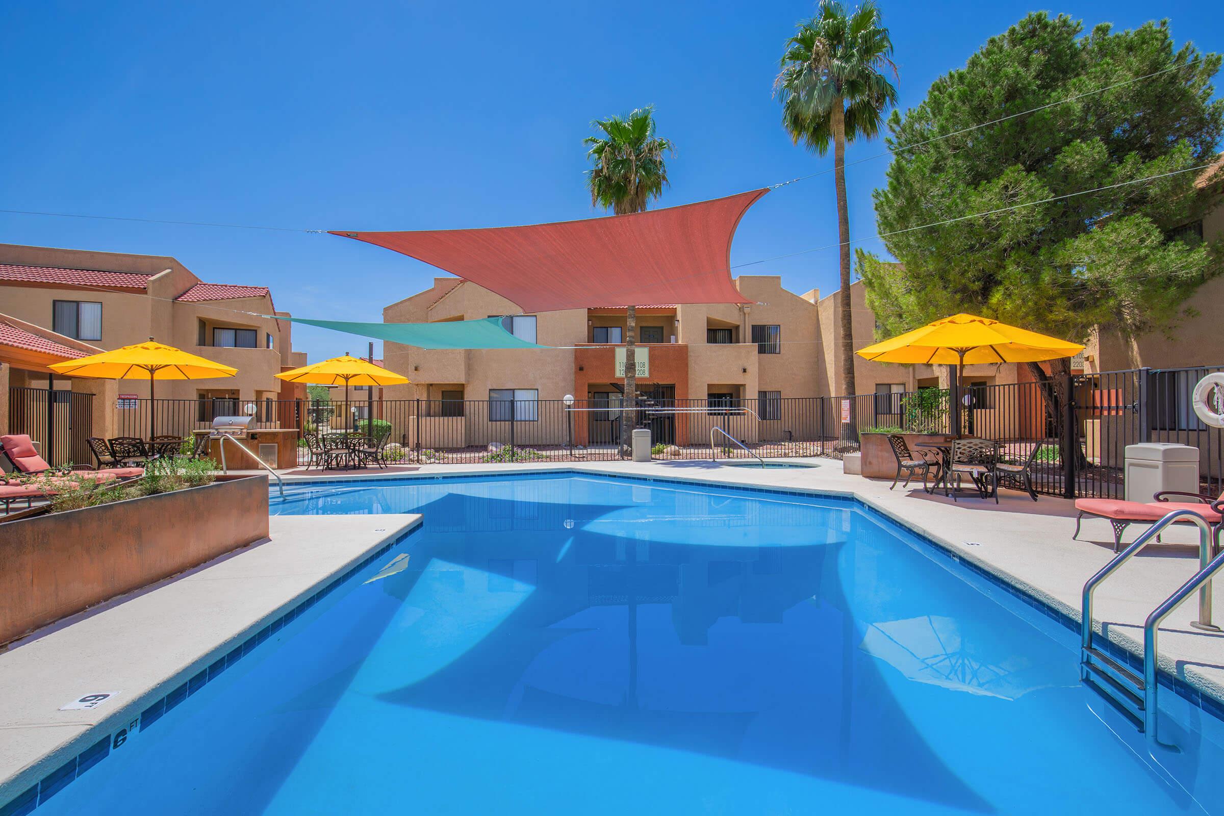 A bright outdoor swimming pool surrounded by lounge chairs and umbrellas. Palm trees are visible in the background, and colorful shade sails are stretched overhead. The pool area is enclosed with a fence, and adjacent buildings can be seen behind the pool. The sky is clear and blue, creating a sunny atmosphere.