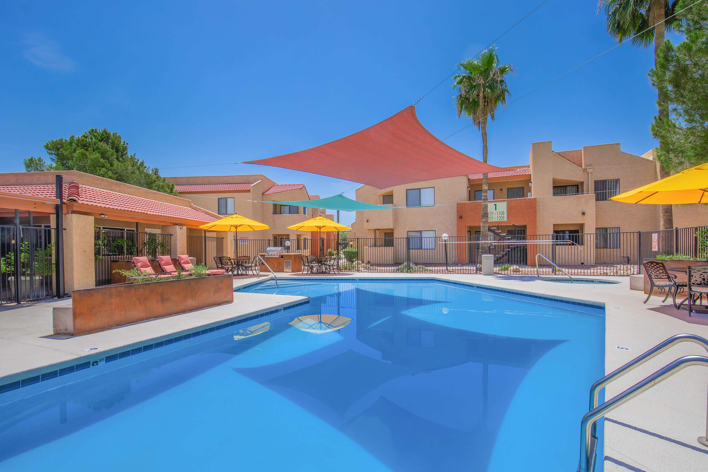 A vibrant pool area featuring crystal-clear water, surrounded by lounge chairs and bright yellow umbrellas. In the background, there are several buildings with peach-colored walls and red roofs, along with lush palm trees and a clear blue sky enhancing the inviting atmosphere.