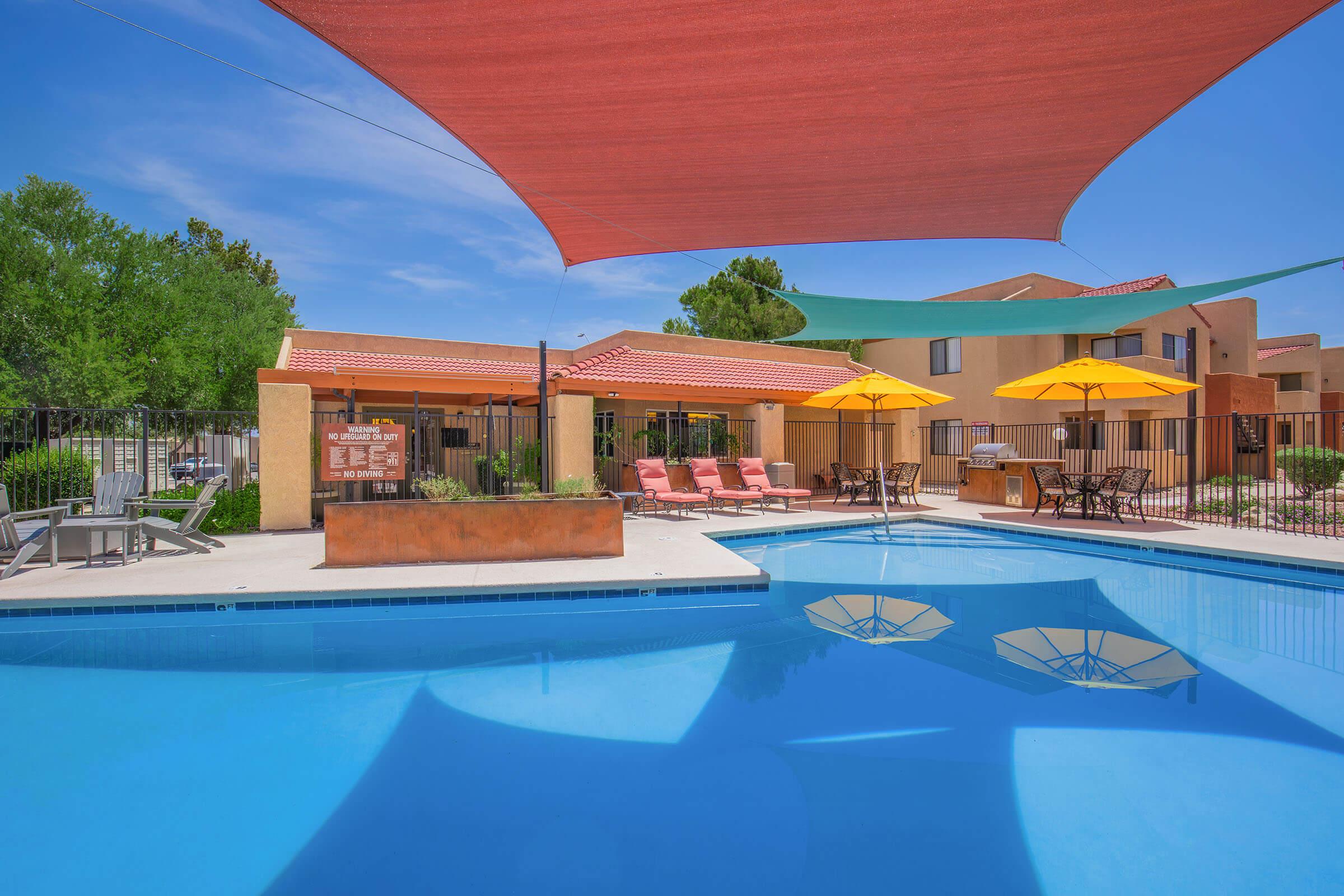 A bright swimming pool with shaded areas, surrounded by lounge chairs and orange umbrellas. In the background, a building with a red roof and lush greenery can be seen. Clear blue sky enhances the tranquil atmosphere. Ideal for relaxation or leisure activities.