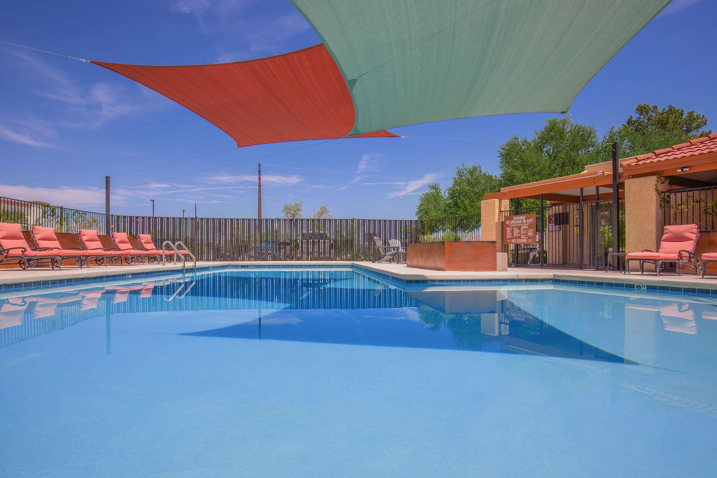 A sunny outdoor pool area featuring a clear blue pool with vibrant red and green shade sails above. Lounge chairs in orange line the pool's edge, and a backdrop of greenery and a decorative wall add to the inviting atmosphere. The sky is bright with a few clouds, indicating a pleasant day.