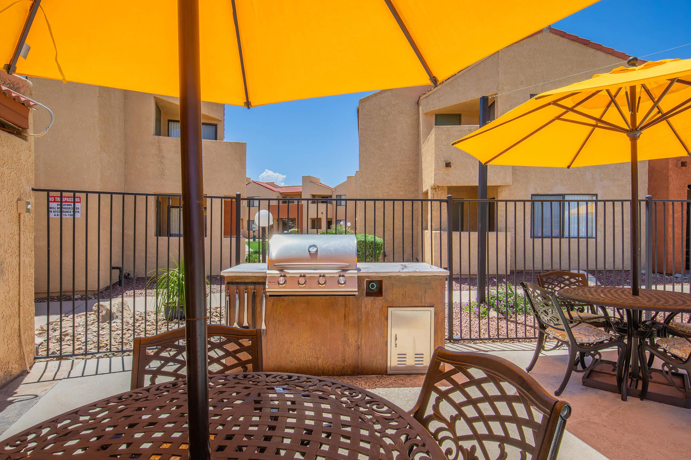 Outdoor grilling area with a stainless steel gas grill beneath a bright yellow umbrella. Surrounding the grill are metal tables with chairs, and in the background, there are beige apartment buildings and a fence surrounding the space. The sky is clear and blue, indicating a sunny day.