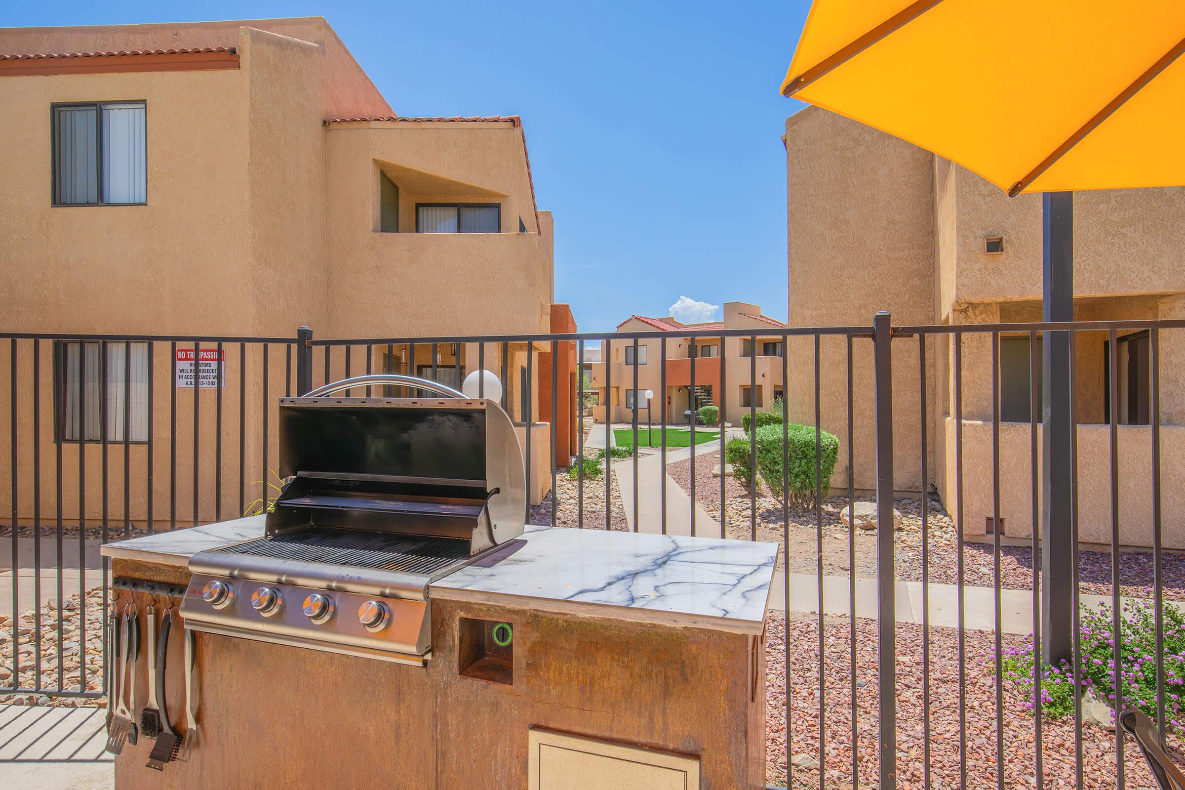 Outdoor patio area featuring a barbecue grill in front of a fenced residential complex. The scene includes two buildings, a clear blue sky, and a yellow umbrella providing shade for the grill area. Decorative stones and green shrubs line the pathways between the buildings.