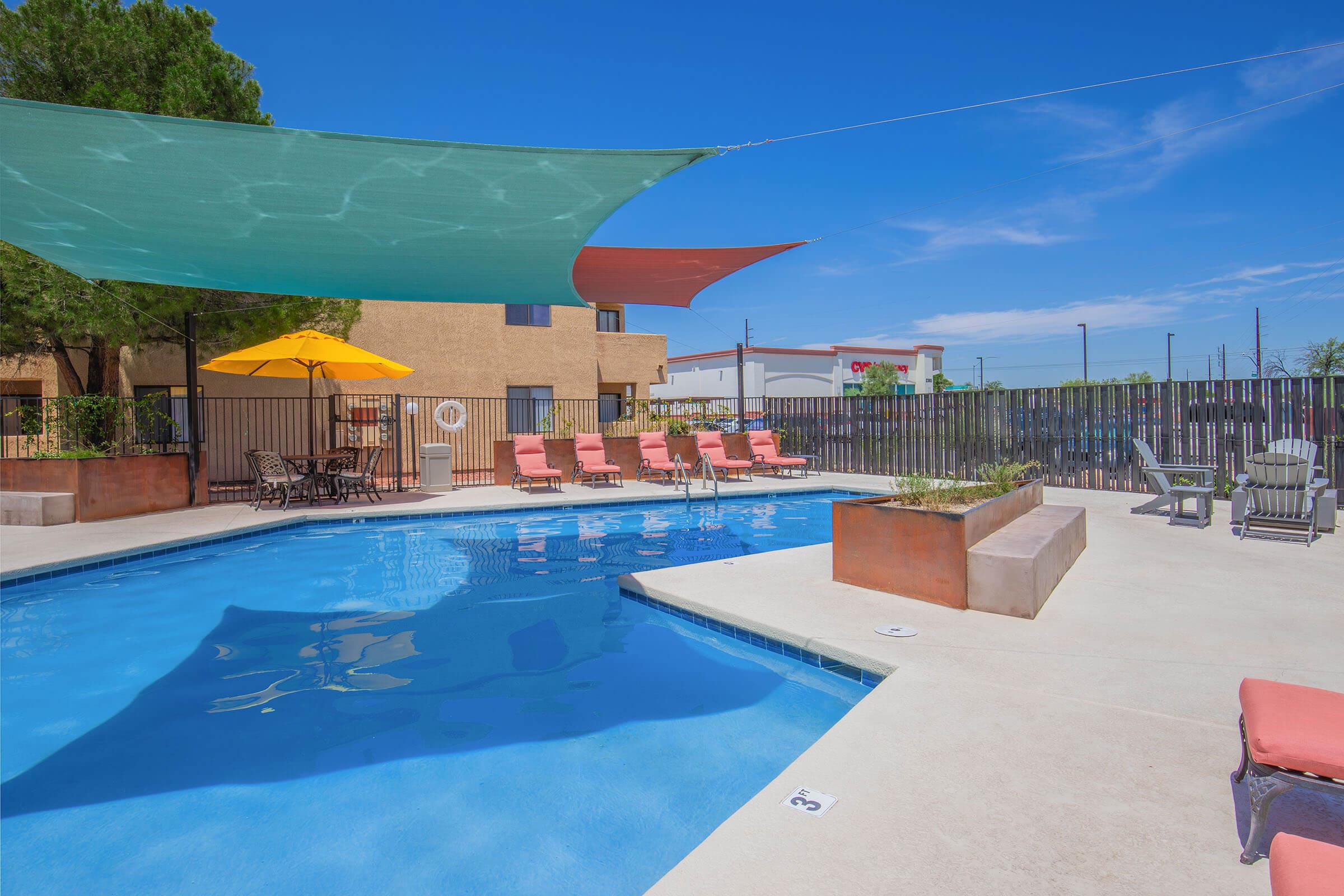 A vibrant outdoor swimming pool surrounded by lounge chairs and shaded by colorful canopies. There is seating with an umbrella, a small landscaped area, and a clear blue sky in the background, creating a relaxing summer atmosphere.