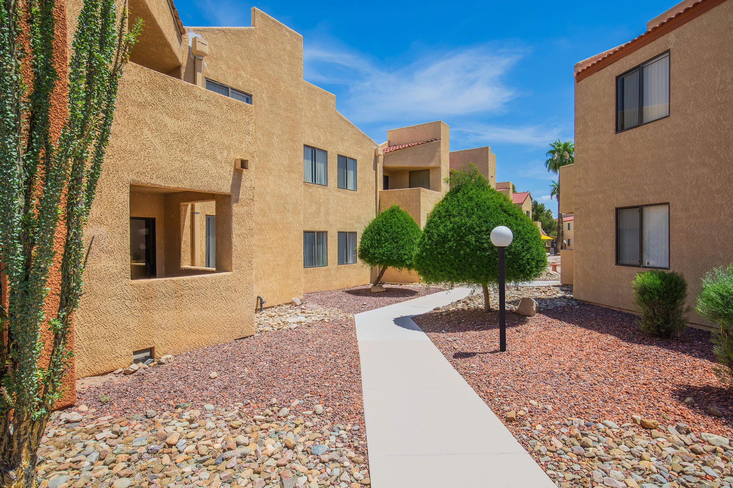 A landscaped pathway meanders through a residential area featuring beige, stucco apartment buildings. Lush green shrubs and small trees frame the walkways, while decorative rocks add texture to the ground. Bright blue skies create a cheerful atmosphere.