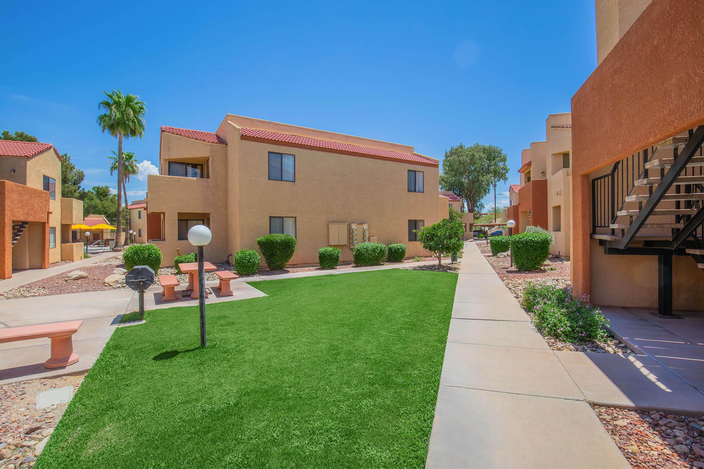 A well-maintained residential complex featuring multiple two-story buildings with beige and orange exteriors. The landscape includes manicured green lawns, decorative bushes, and palm trees. Walkways connect the buildings, and there are outdoor seating areas with lamps along the paths under a clear blue sky.