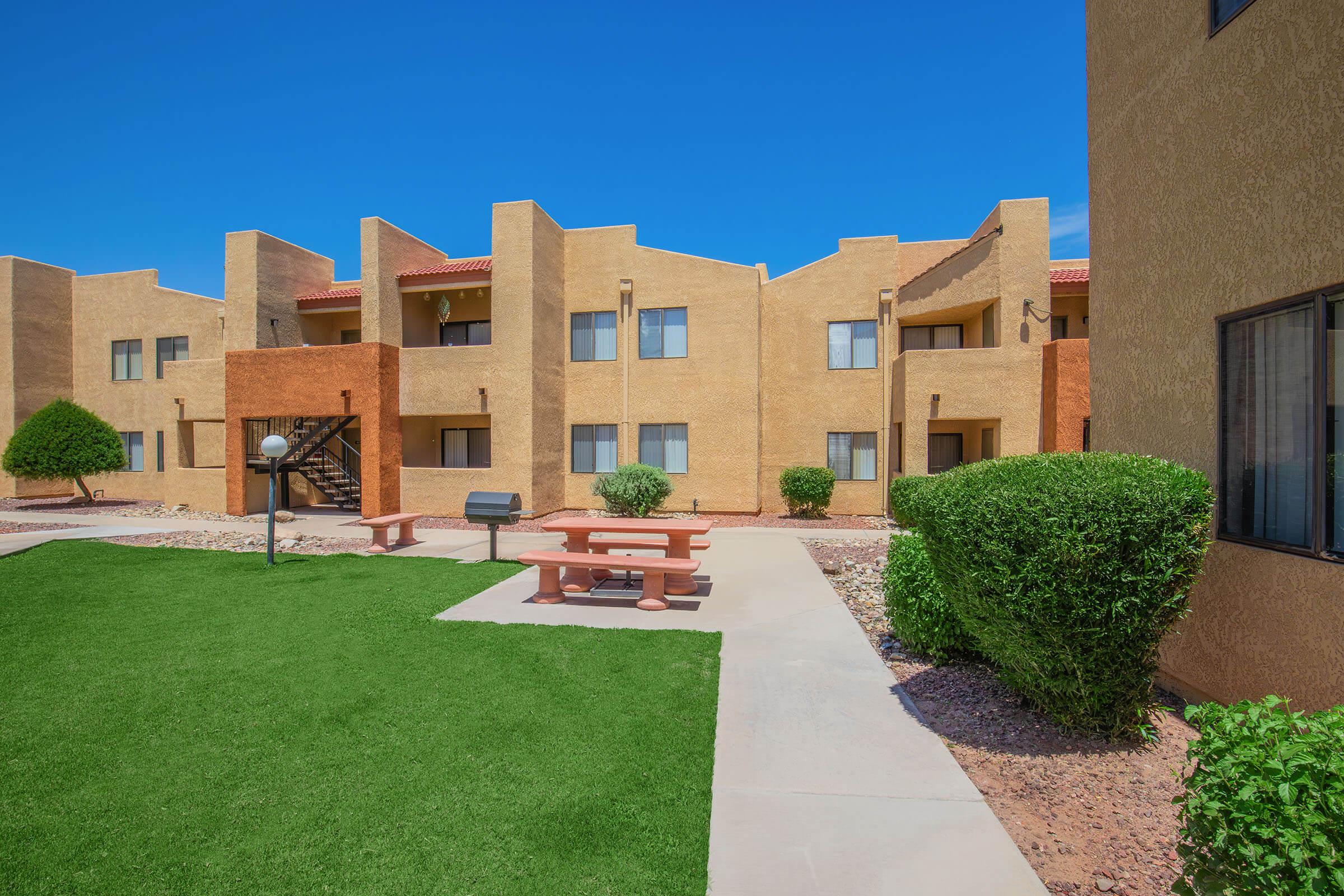View of a landscaped courtyard featuring a grassy area, picnic table, and barbecue grill surrounded by beige apartment buildings under a clear blue sky. The setting reflects a well-maintained apartment complex with multiple units and a sunny atmosphere.